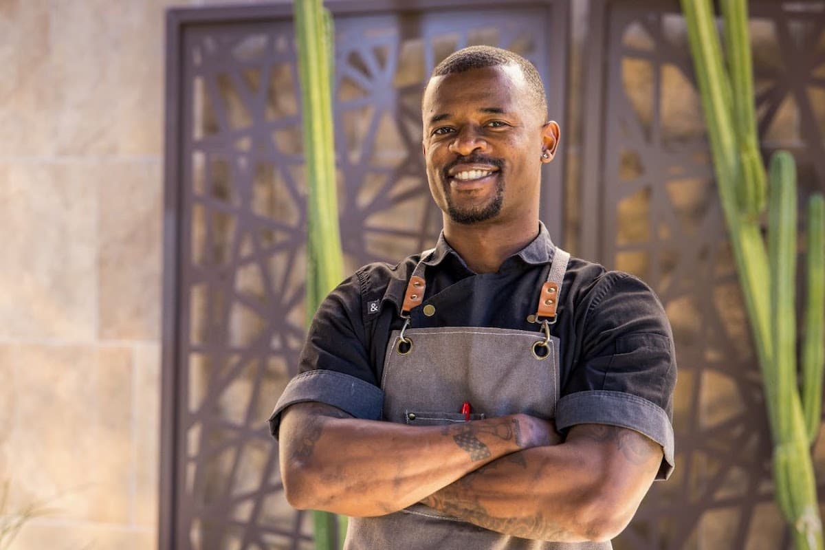 Smiling chef in apron standing confidently with arms crossed in a modern desert setting.