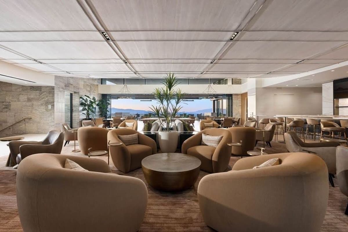 Spacious modern lounge with curved seating, neutral tones, and open views toward a dining area and outdoor landscape.