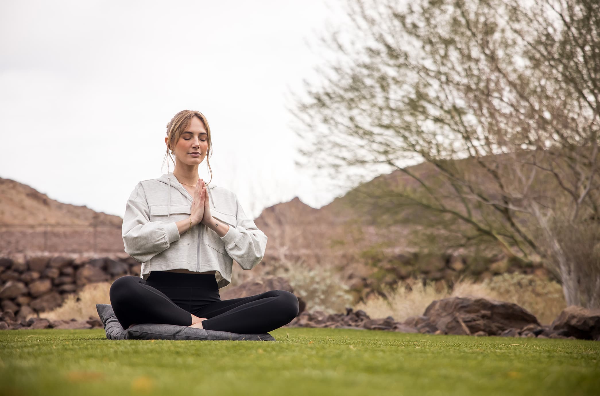 A resident practicing yoga outdoors, seated in meditation on a manicured lawn surrounded by desert landscape, rocks, and mountain views.
