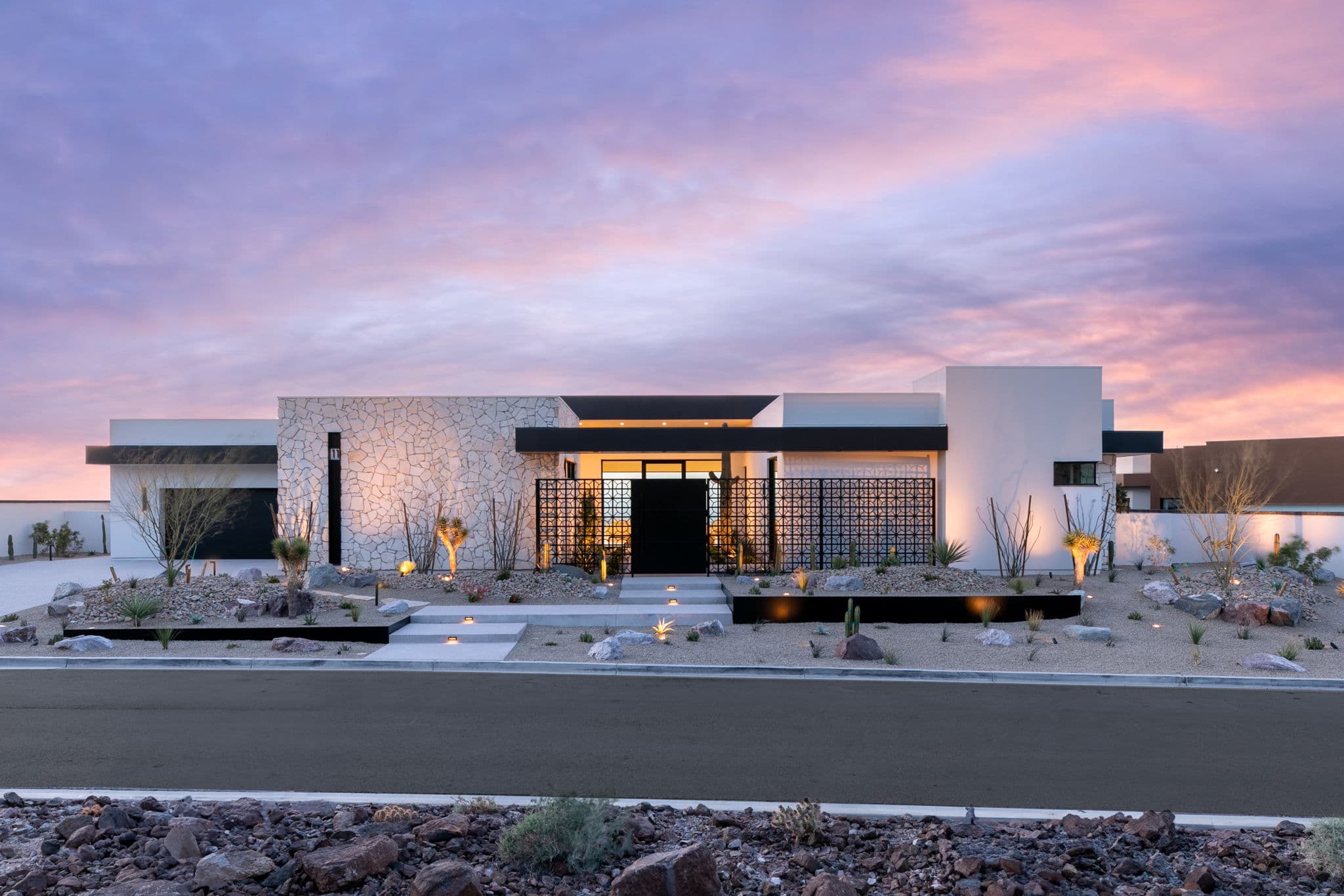 Contemporary desert house with minimalist design, stone facade, landscaped yard, and ambient exterior lighting at sunset.