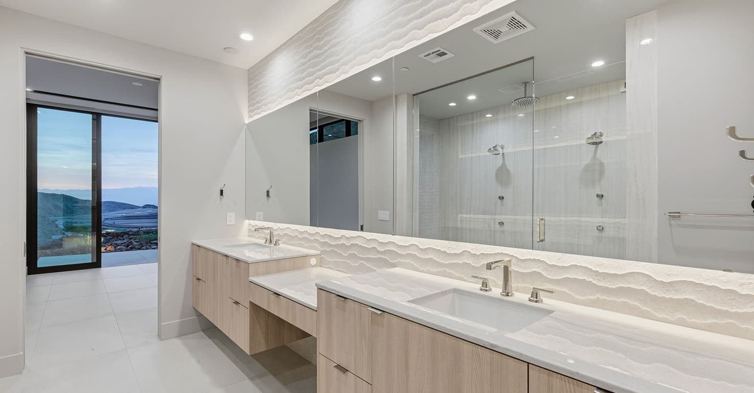 Modern spa-inspired bathroom with dual floating vanities, illuminated textured backsplash, oversized glass walk-in shower, and serene desert views