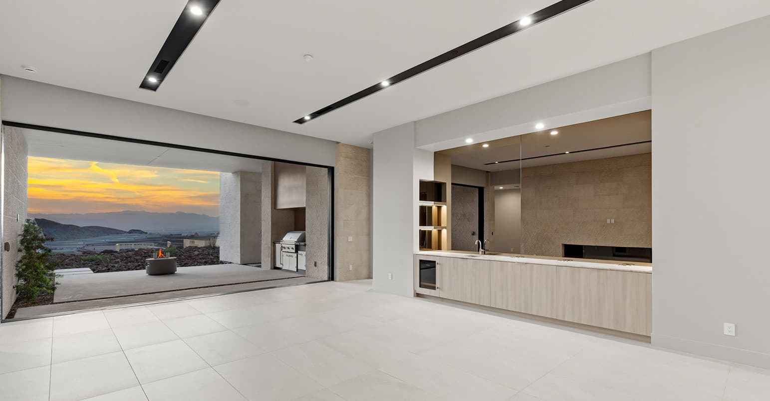 Minimalist indoor-outdoor living space with sleek wet bar, floor-to-ceiling sliding glass walls, and covered terrace featuring a modern fire feature and sunset desert views