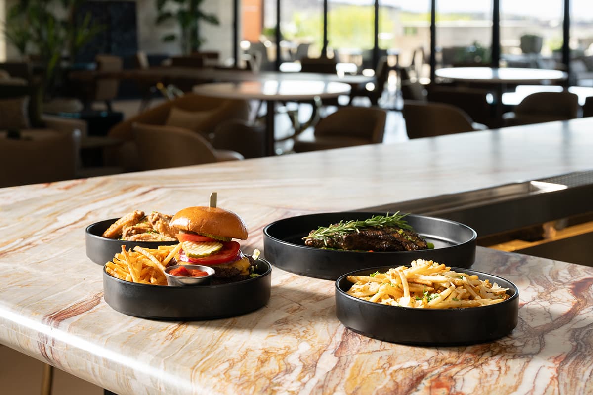 Plates of food including a burger with fries, pasta, and a steak are arranged on a marble bar counter in a modern, softly lit restaurant.
