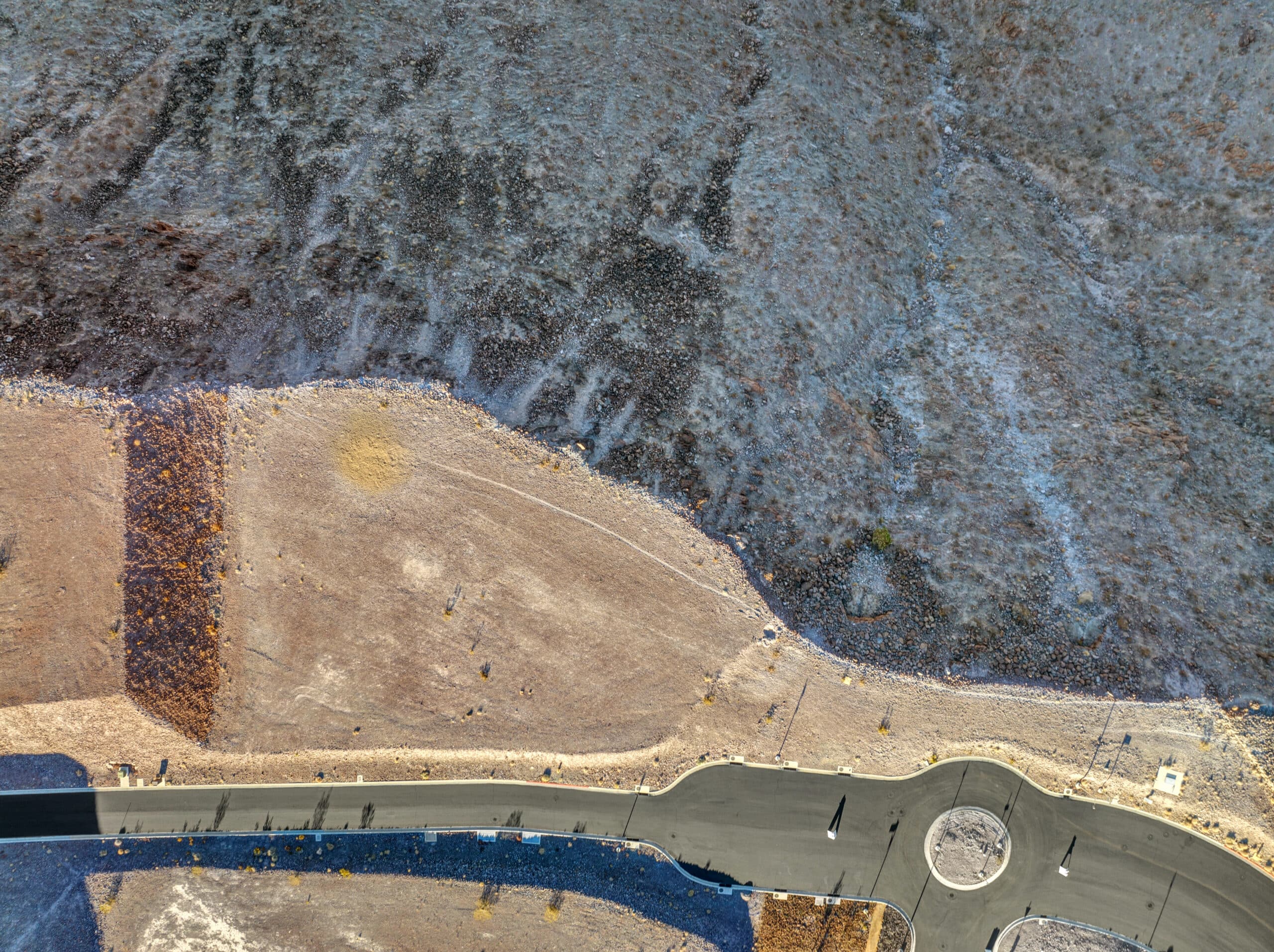 Overhead aerial view of a homesite at Ascaya, showing the building pad positioned along a curved roadway and roundabout, bordered by natural desert terrain and rugged mountainside slopes.