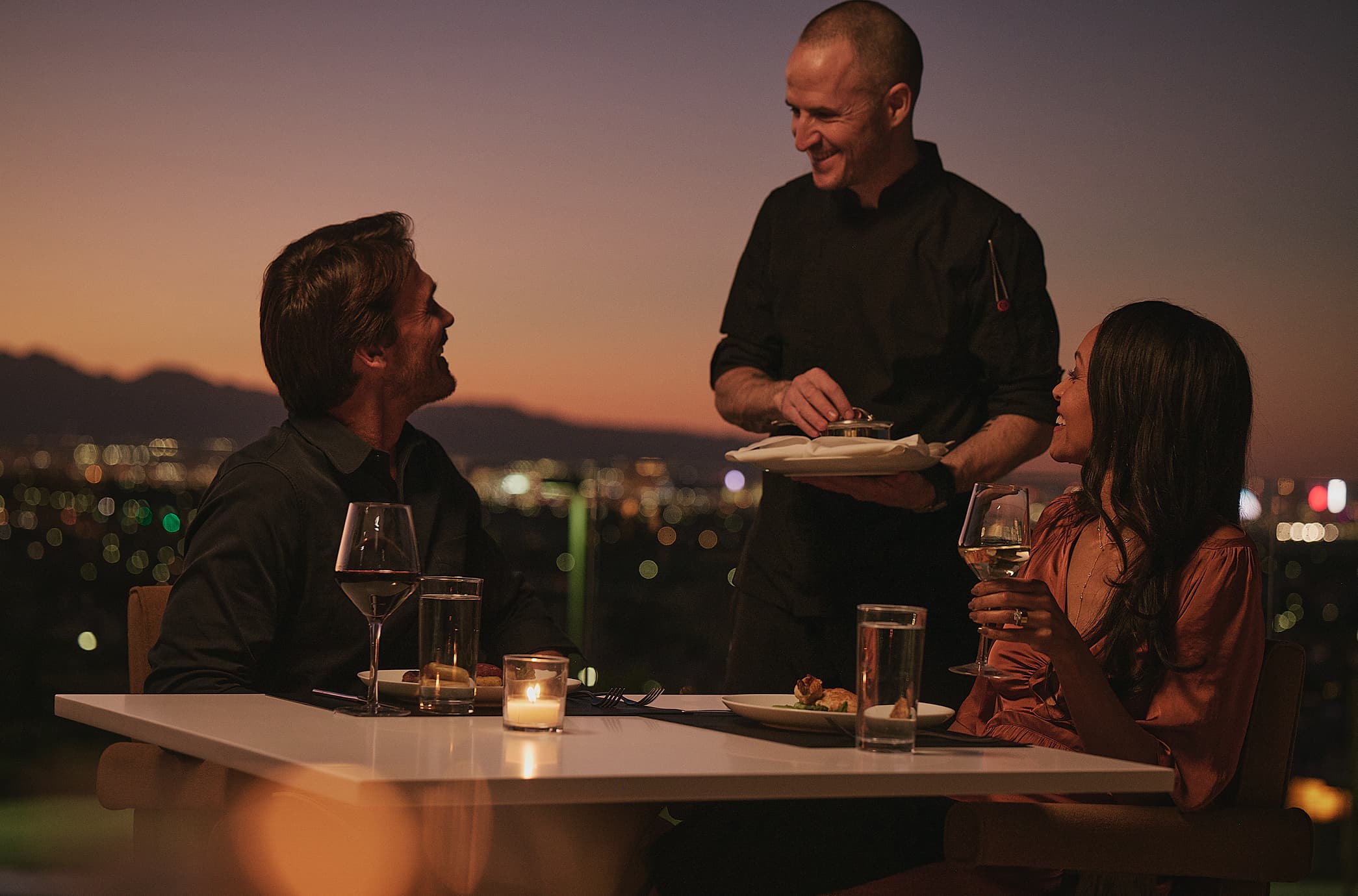 A man and woman sit at an outdoor table at sunset, smiling at each other while holding glasses of wine, with a city skyline glowing in the background.