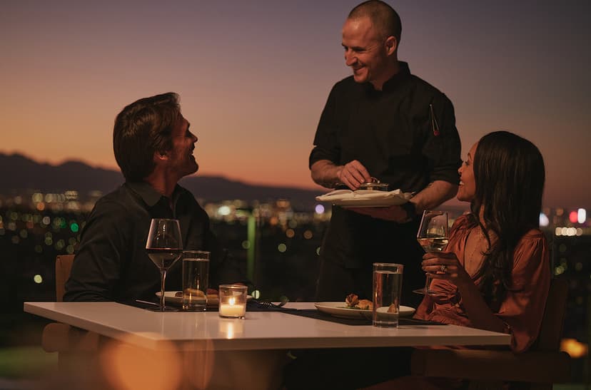 A man and woman sit at an outdoor table at sunset, smiling at each other while holding glasses of wine, with a city skyline glowing in the background.