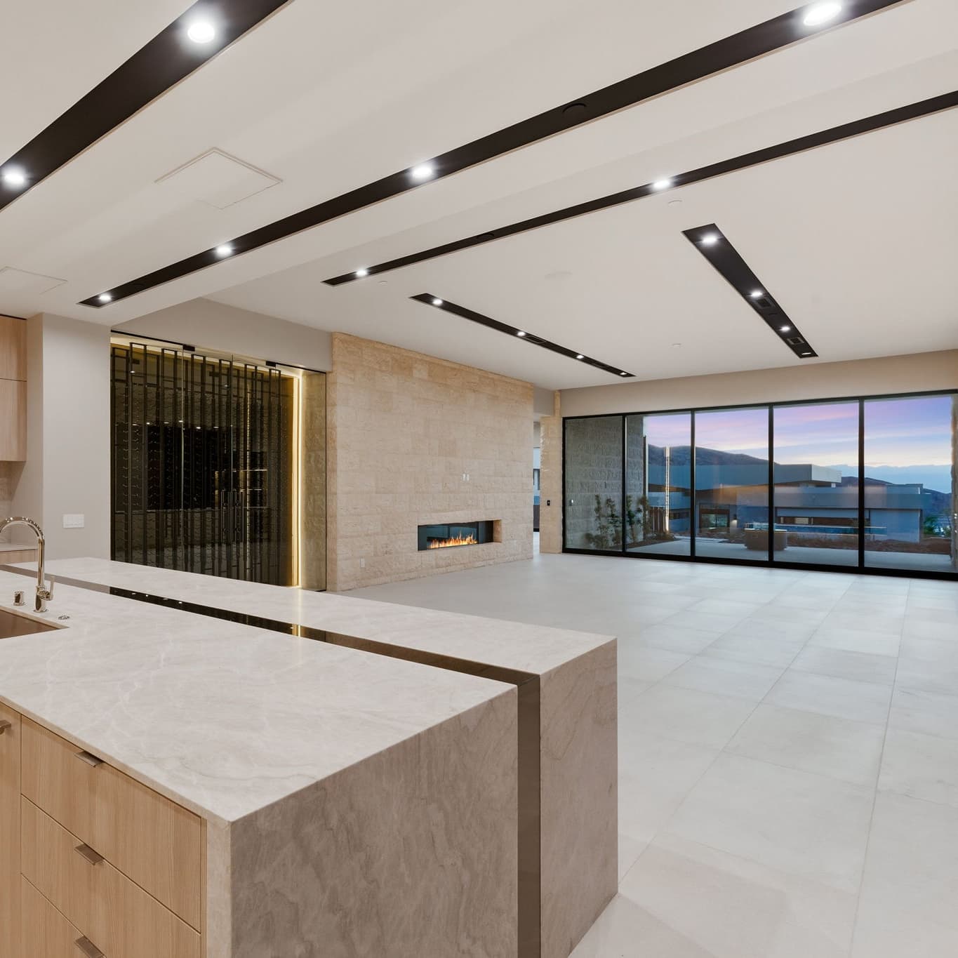 Open-concept great room featuring a chef’s kitchen, wine display, linear fireplace, floor-to-ceiling windows, and seamless indoor-outdoor living