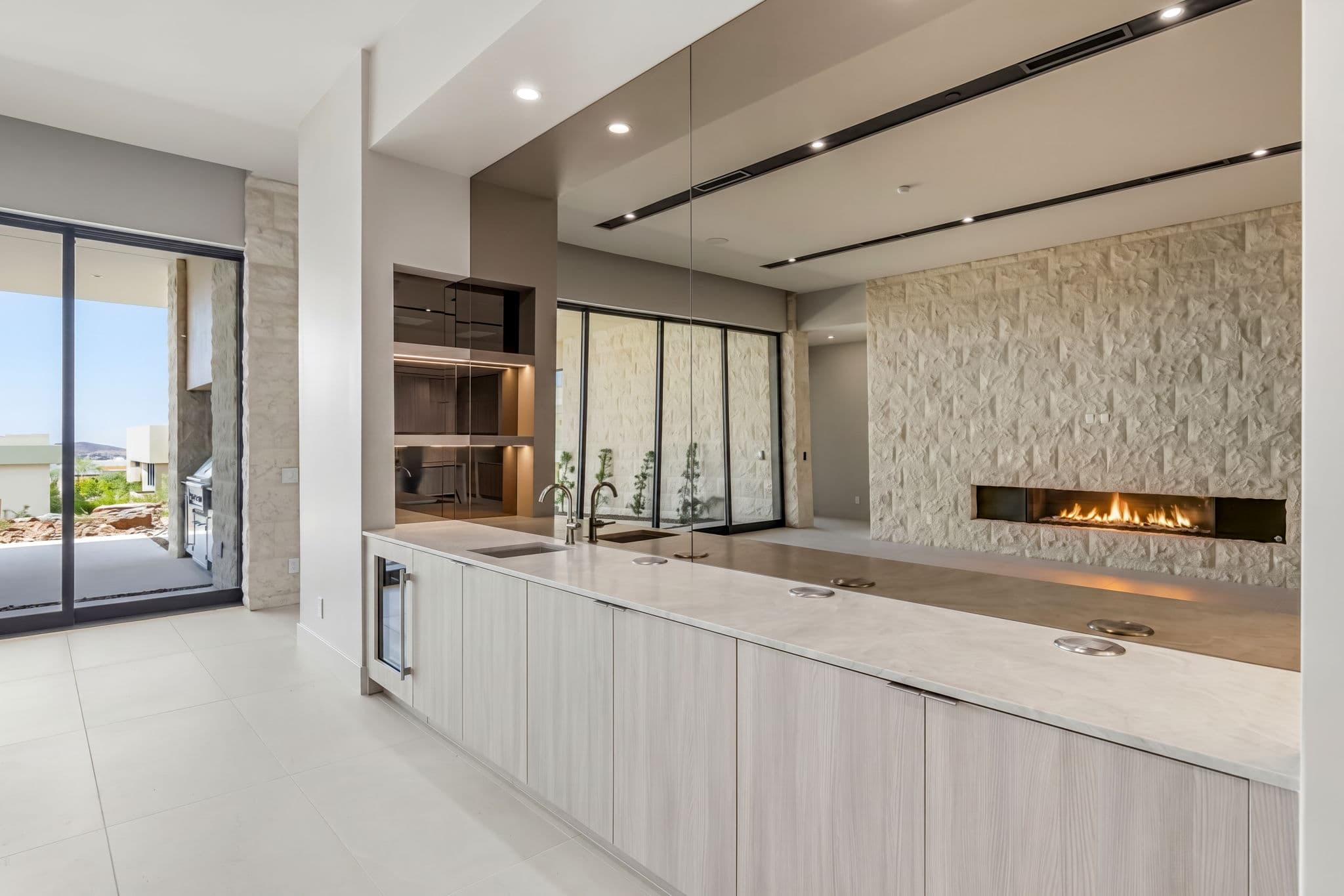 Contemporary wet bar with waterfall-edge countertop, built-in beverage refrigerator, custom cabinetry, and a linear fireplace framed by textured stone