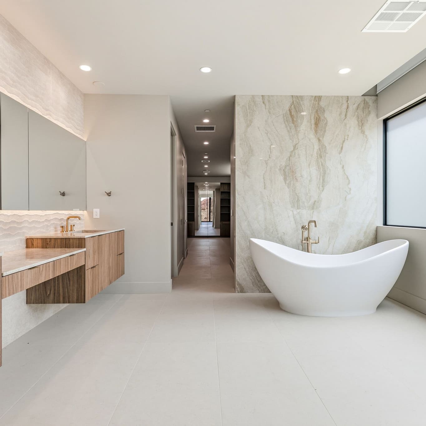 Spa-inspired primary bathroom with freestanding soaking tub, floating wood vanity, illuminated mirror, and floor-to-ceiling stone accent wall