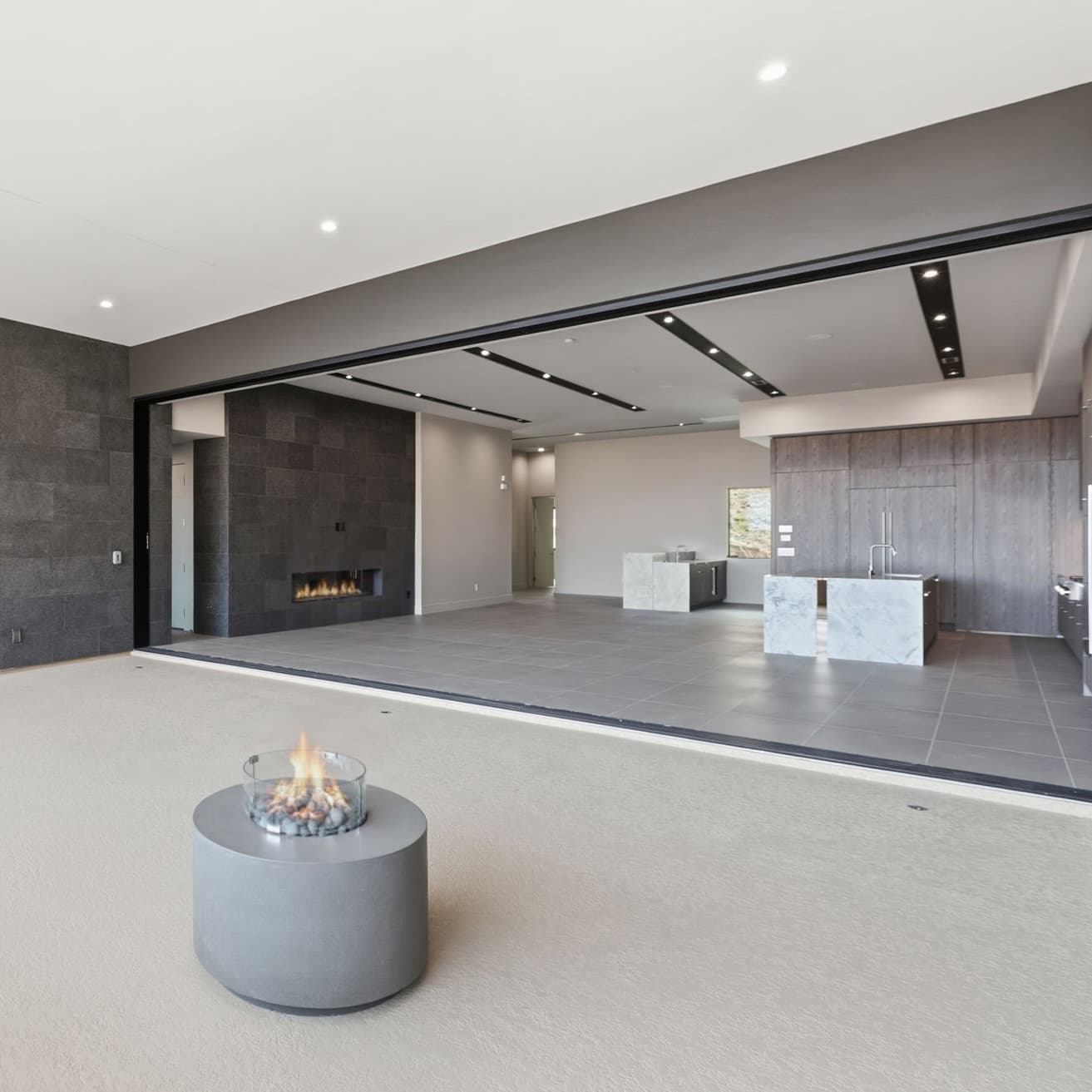 Modern indoor-outdoor living area with retractable glass wall, dual fireplaces, minimalist kitchen, and expansive open-concept design