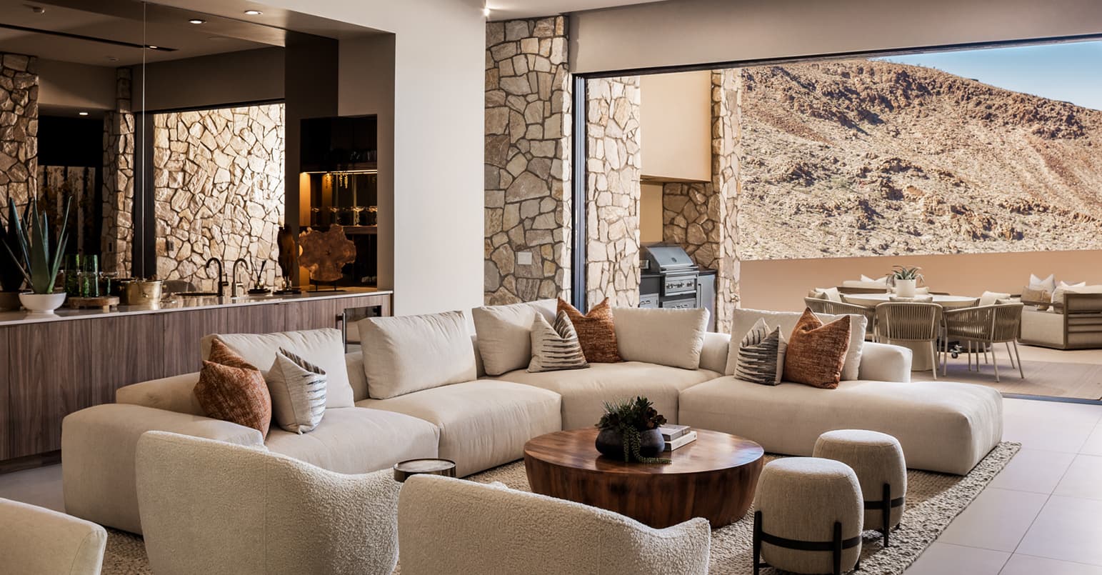 Spacious open-concept living room with a large sectional sofa, warm wood accents, and a built-in bar, seamlessly extending to a covered outdoor terrace with dining and grill, framed by desert mountain views.