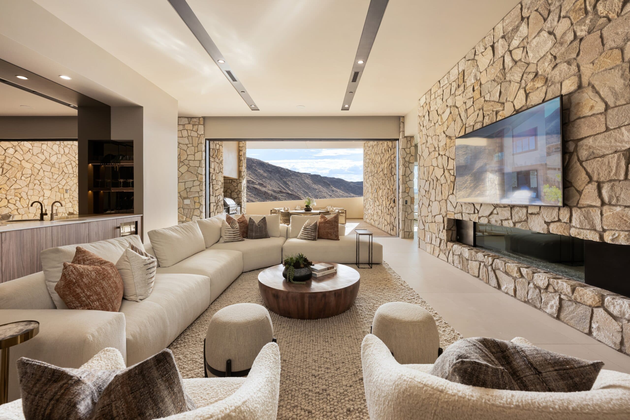 Luxury modern living room with a stone fireplace, neutral furnishings, and seamless indoor-outdoor mountain views.
