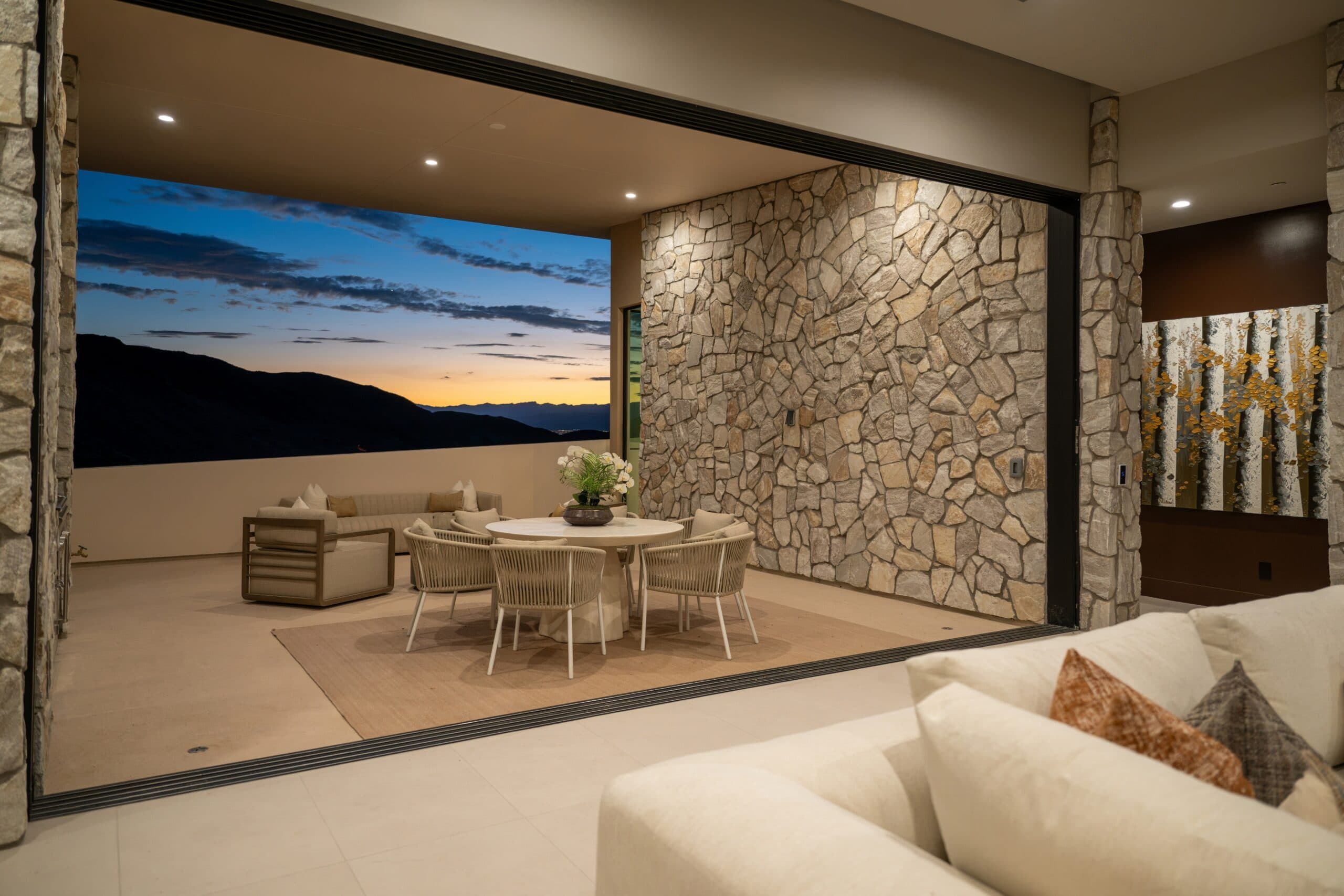 Luxury covered outdoor dining and lounge area with stone walls and panoramic mountain views at sunset.