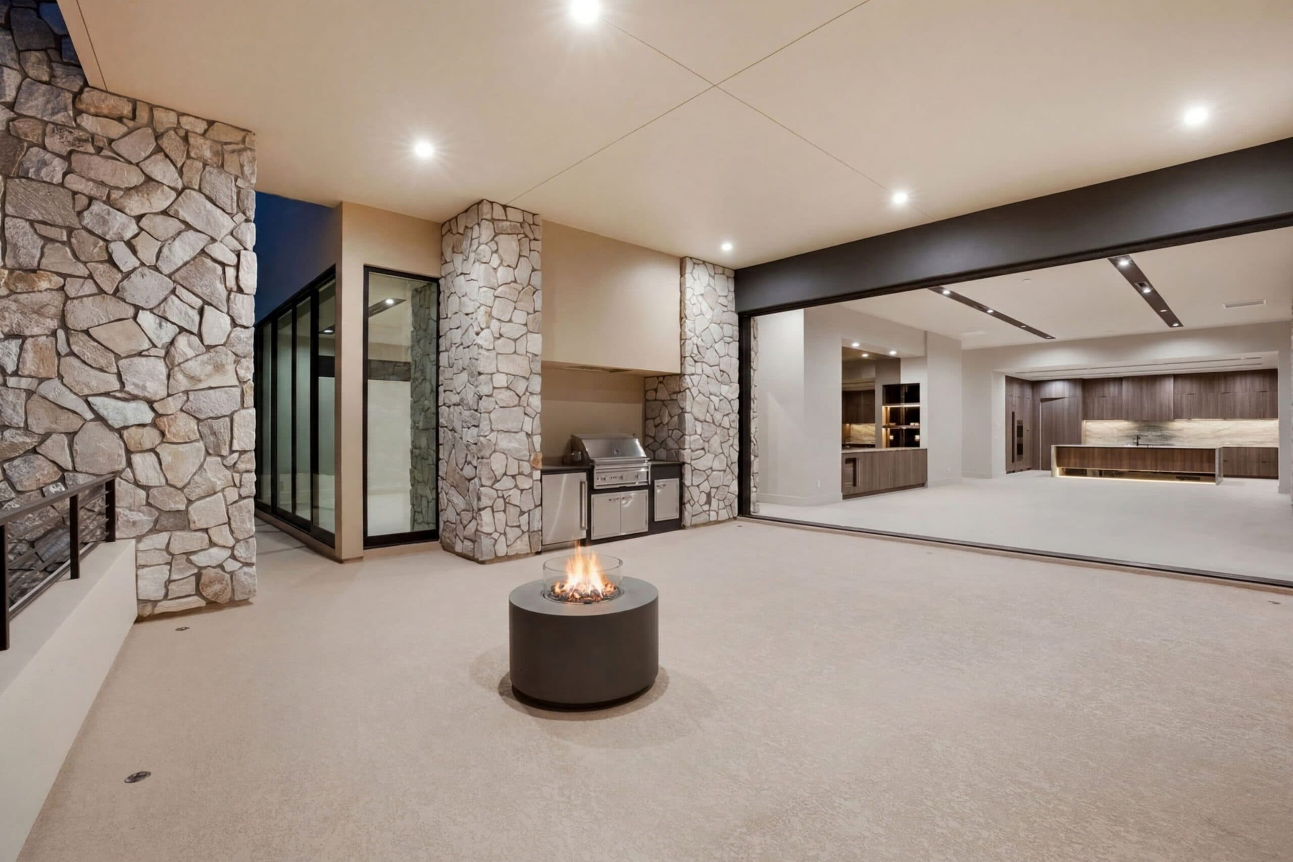 Luxury indoor-outdoor living space with stone columns, a built-in grill, fire pit, and seamless connection to a modern kitchen.