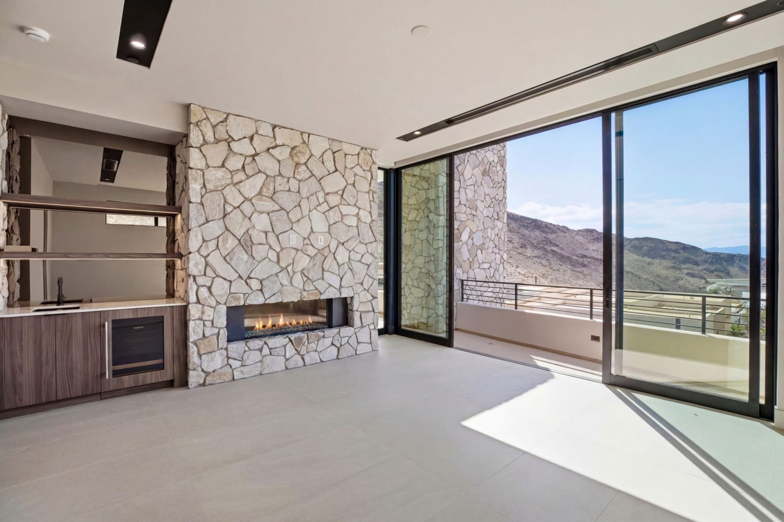 Modern luxury bedroom with a stone fireplace, built-in wet bar, floor-to-ceiling glass doors, and panoramic mountain views.