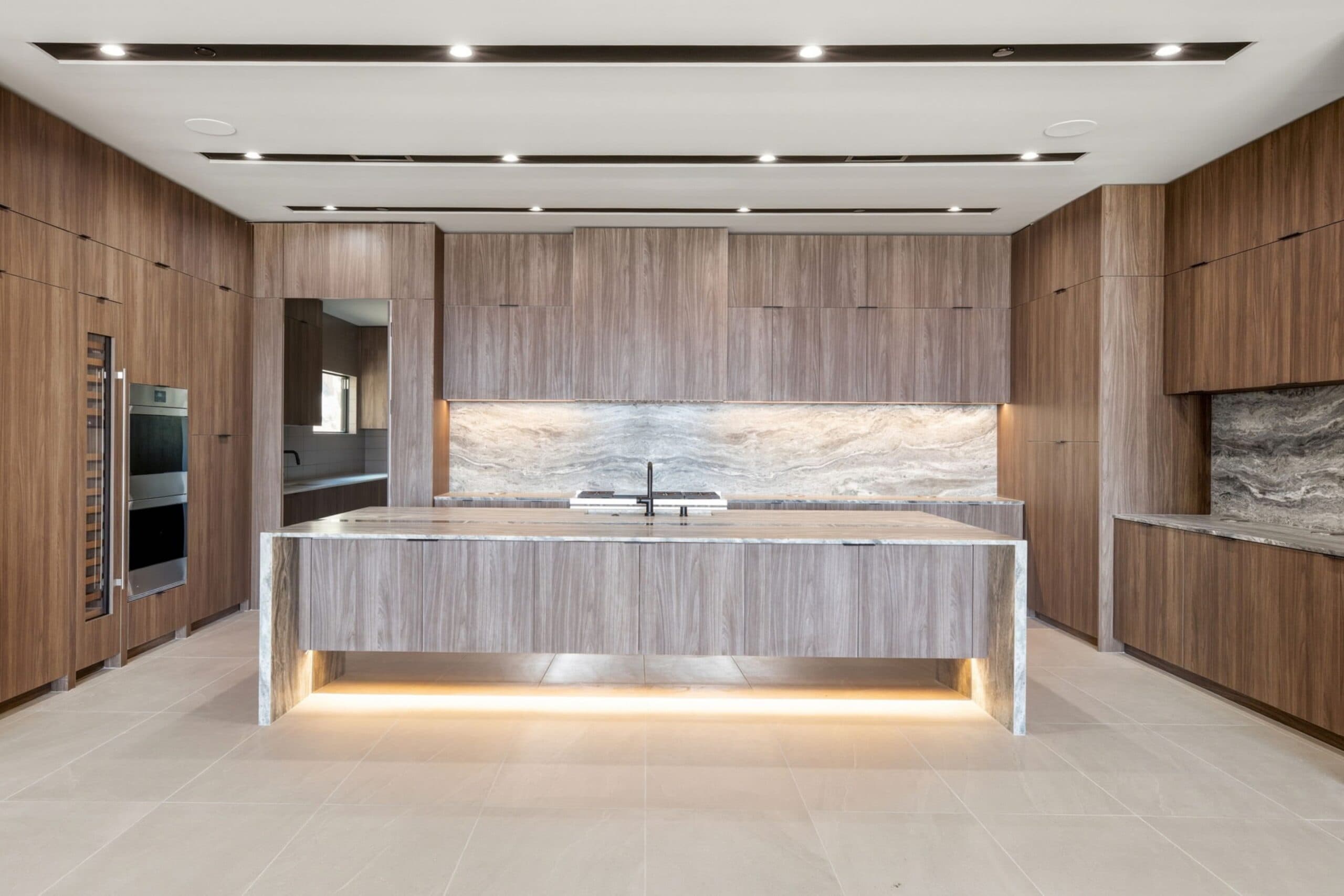 Modern luxury kitchen with floor-to-ceiling wood cabinetry, a large illuminated island, stone backsplash, and sleek integrated appliances.