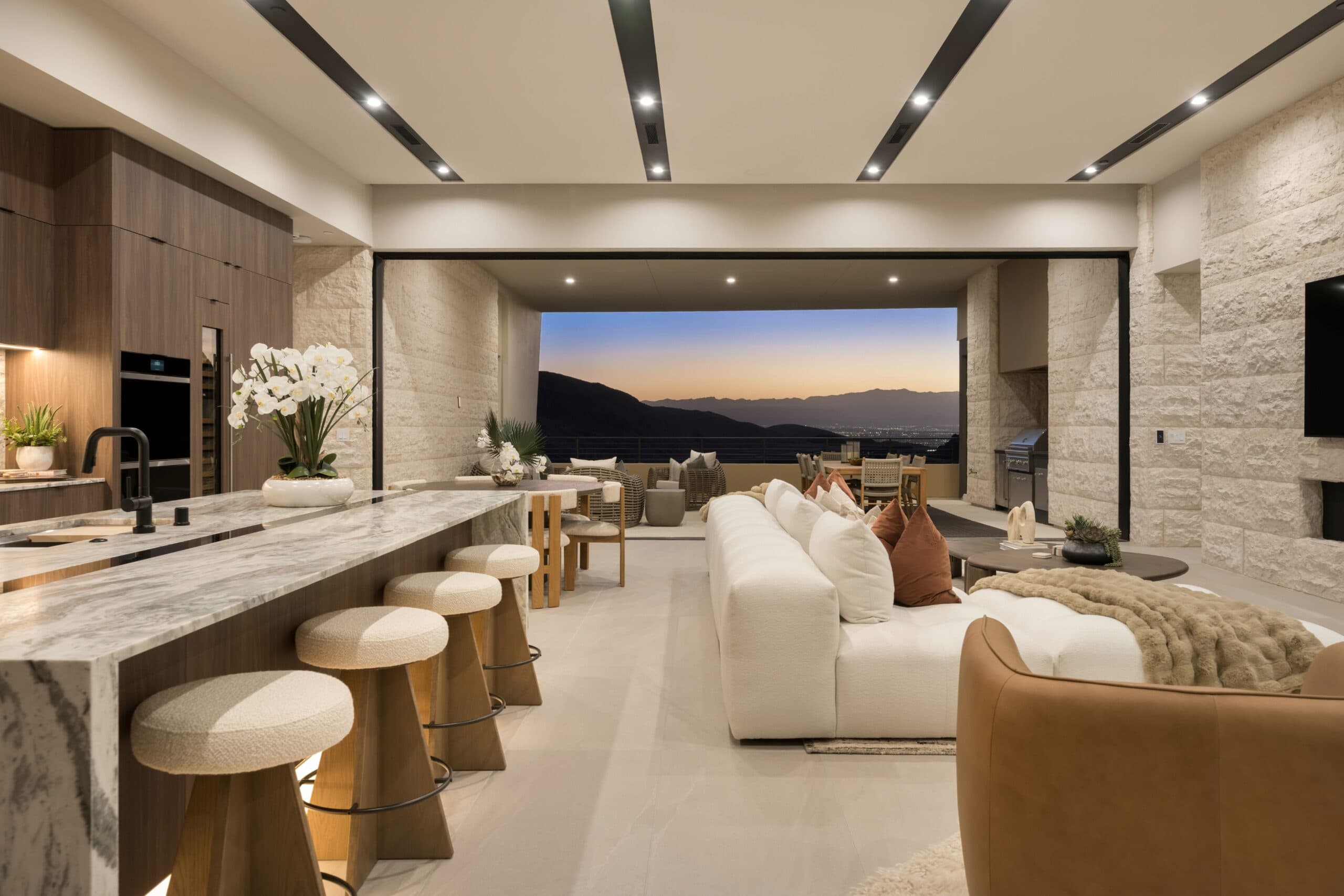 Luxury open-concept kitchen and living space with marble finishes, designer furnishings, seamless indoor-outdoor living, and panoramic mountain views at sunset.