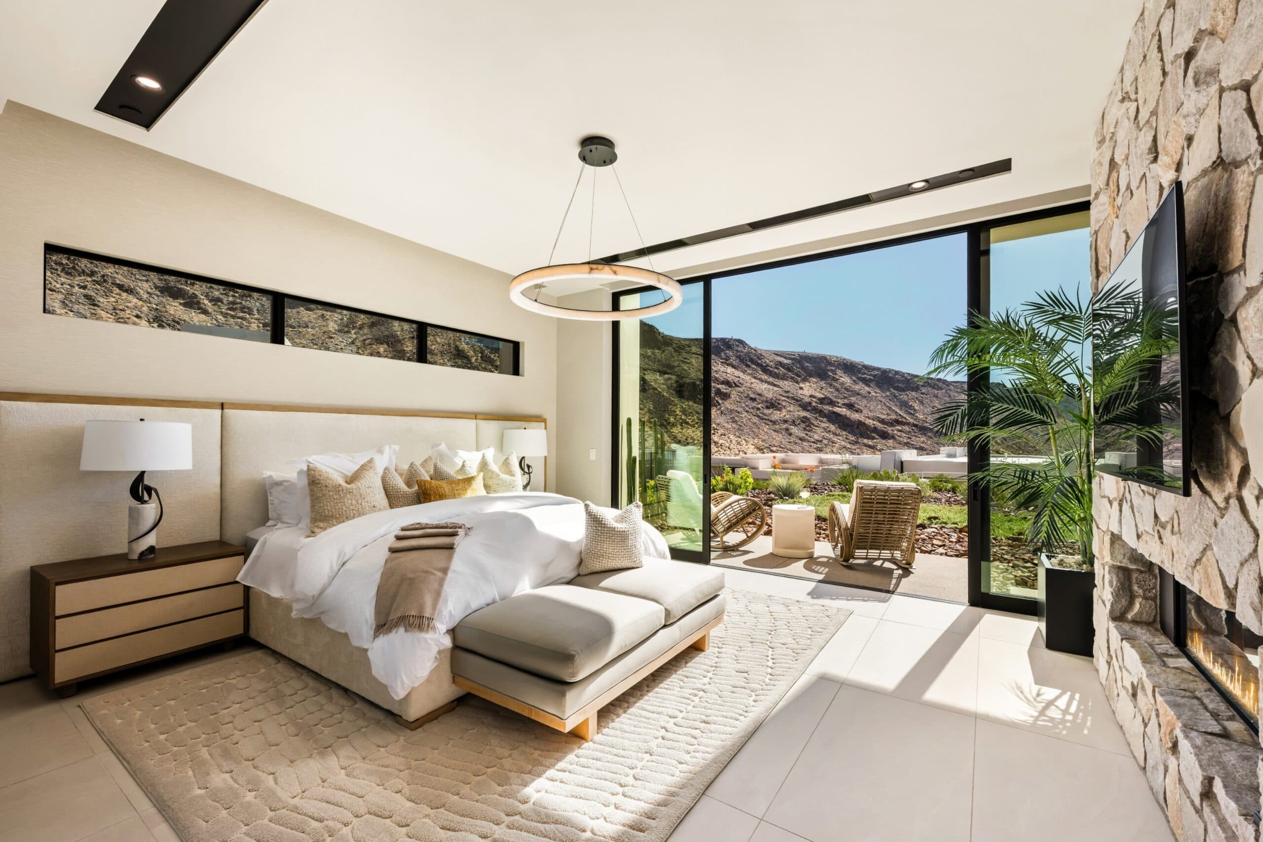 Luxury modern bedroom with floor-to-ceiling glass doors, a stone fireplace, and panoramic desert mountain views.
