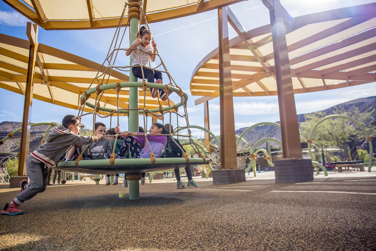 Children playing on a modern playground with climbing structure, shaded canopy, and scenic mountain backdrop.