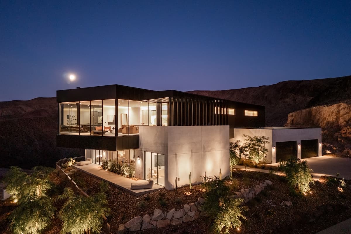 Modern hillside home with expansive glass walls glowing at dusk under a full moon.