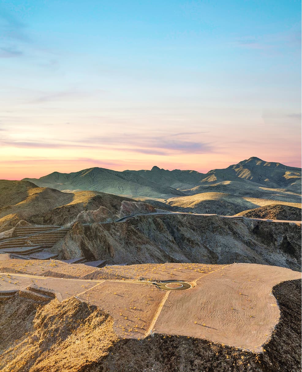 Aerial view of a desert hillside lot with a circular driveway pad overlooking a sprawling suburban city at sunset, with mountains in the distance.