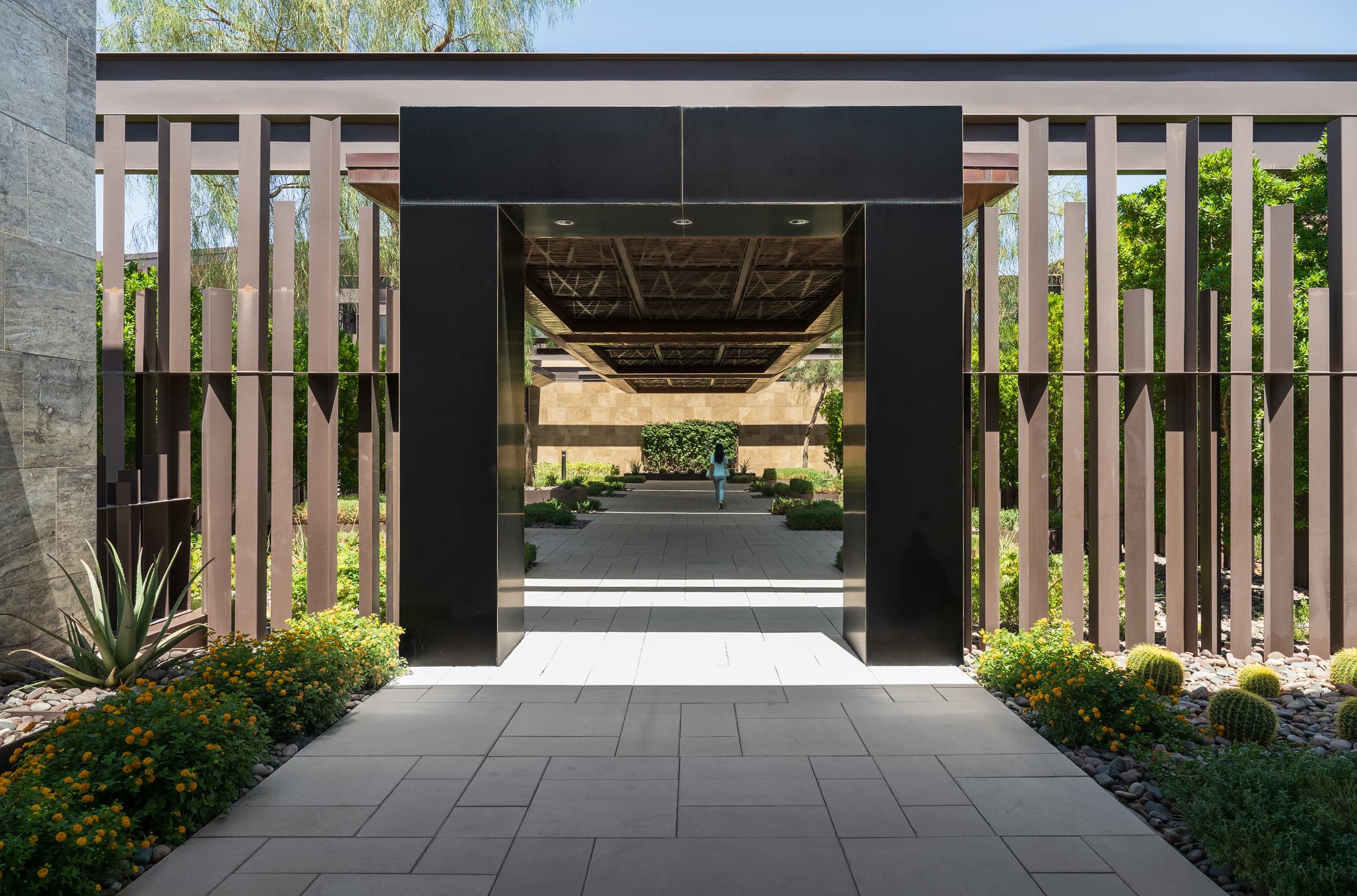 Modern luxury community entrance with sleek architectural gate, landscaped walkway, and desert greenery.