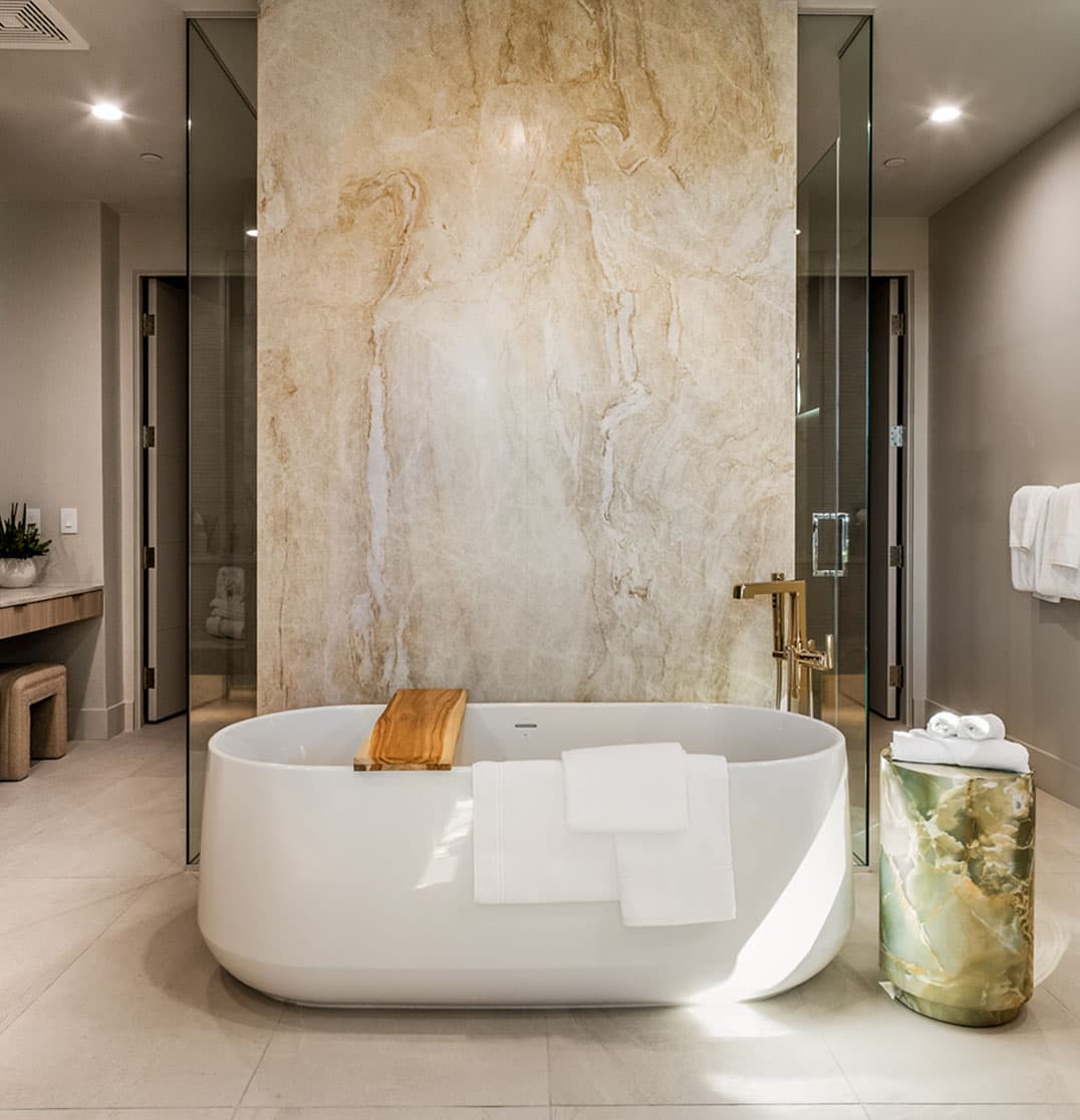 Luxury spa-style bathroom with freestanding soaking tub, floor-to-ceiling stone feature wall, glass shower, and modern gold fixtures.