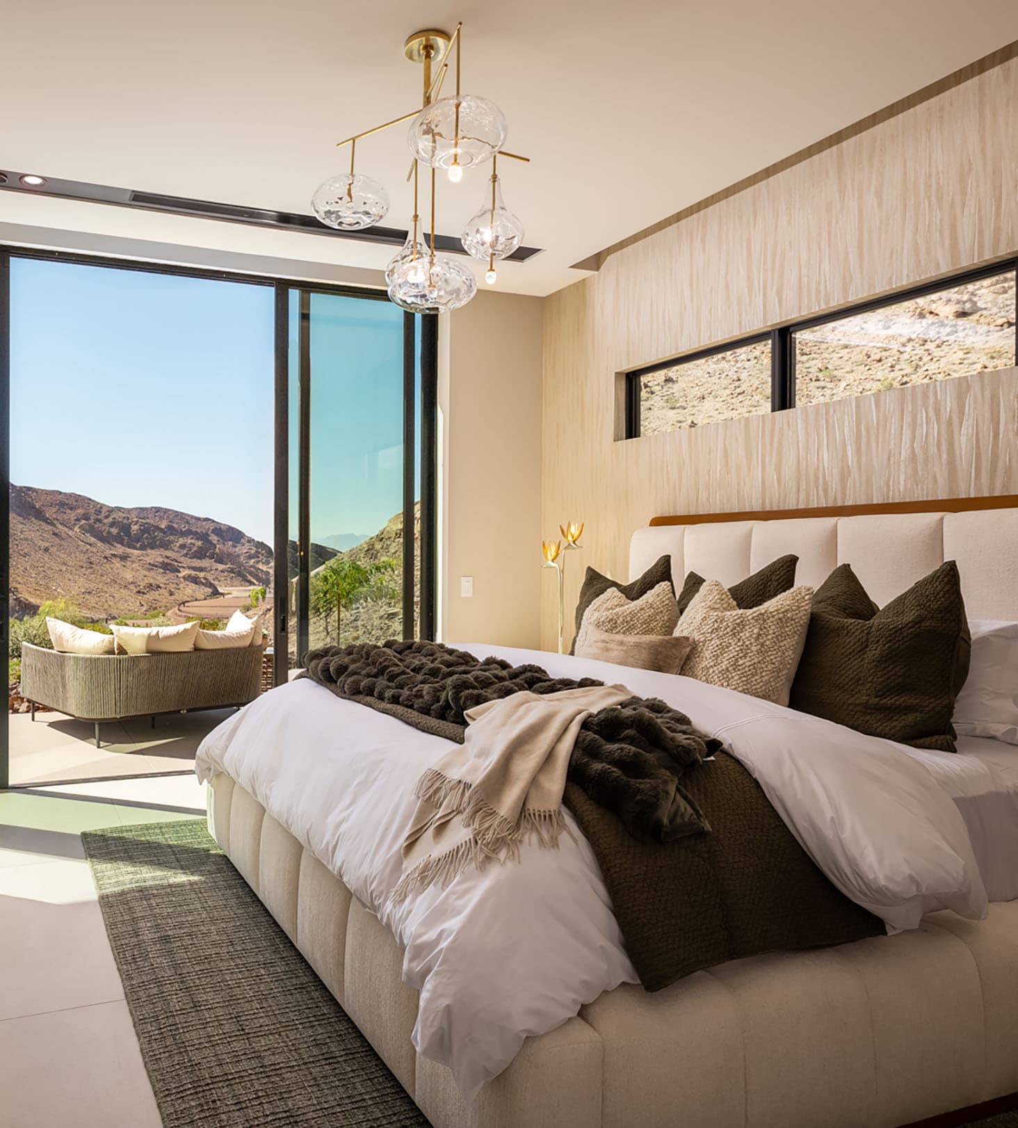 Modern luxury bedroom with plush king bed, designer lighting, and floor-to-ceiling glass doors opening to a scenic desert mountain view patio.