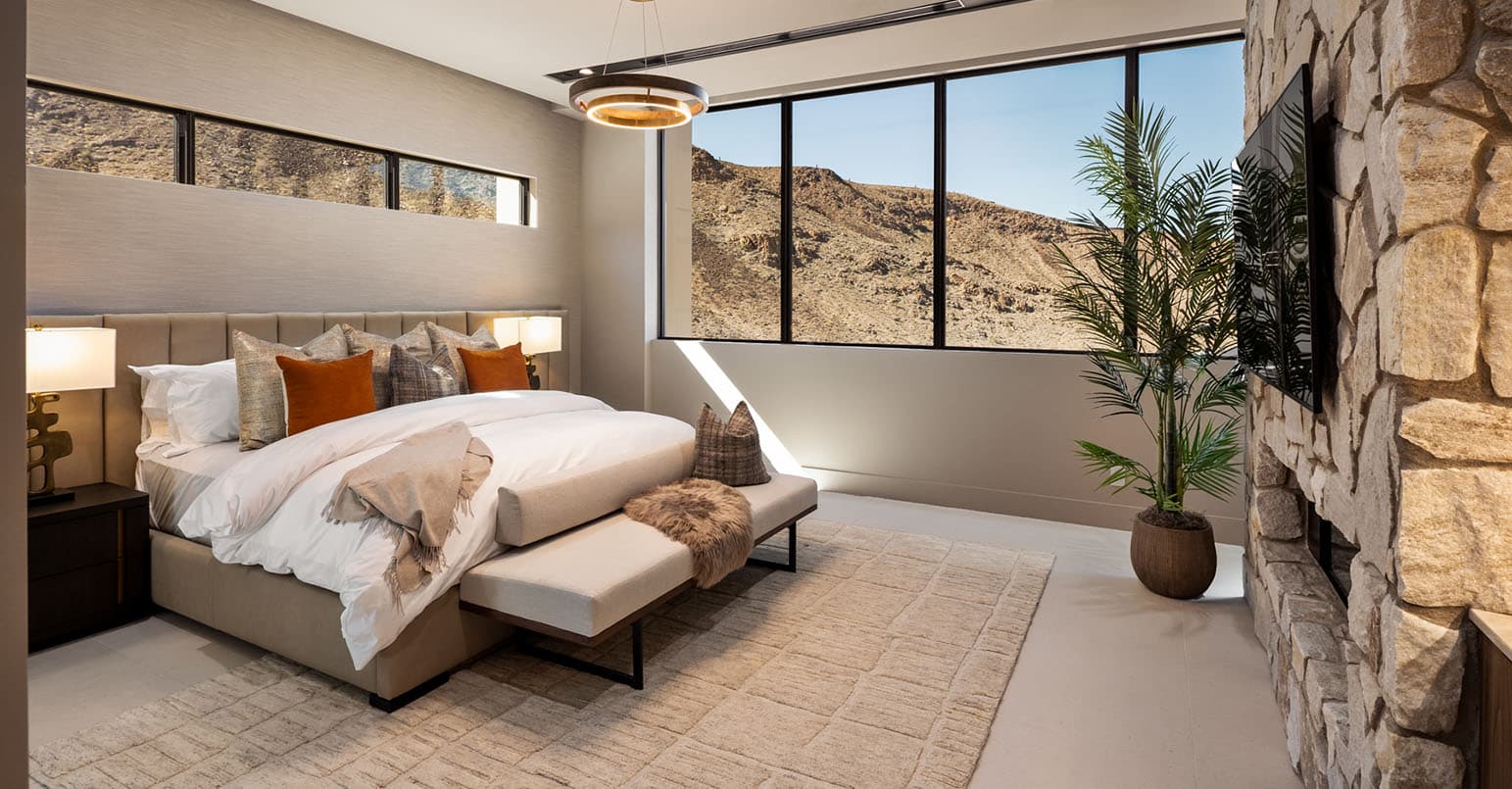 Modern luxury bedroom with upholstered bed, stone accent wall, large windows showcasing desert mountain views, and contemporary decor.