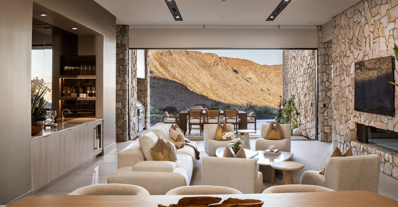 Modern luxury living room with stone accent walls, open-concept design, and seamless indoor-outdoor flow to a desert view patio with dining area.