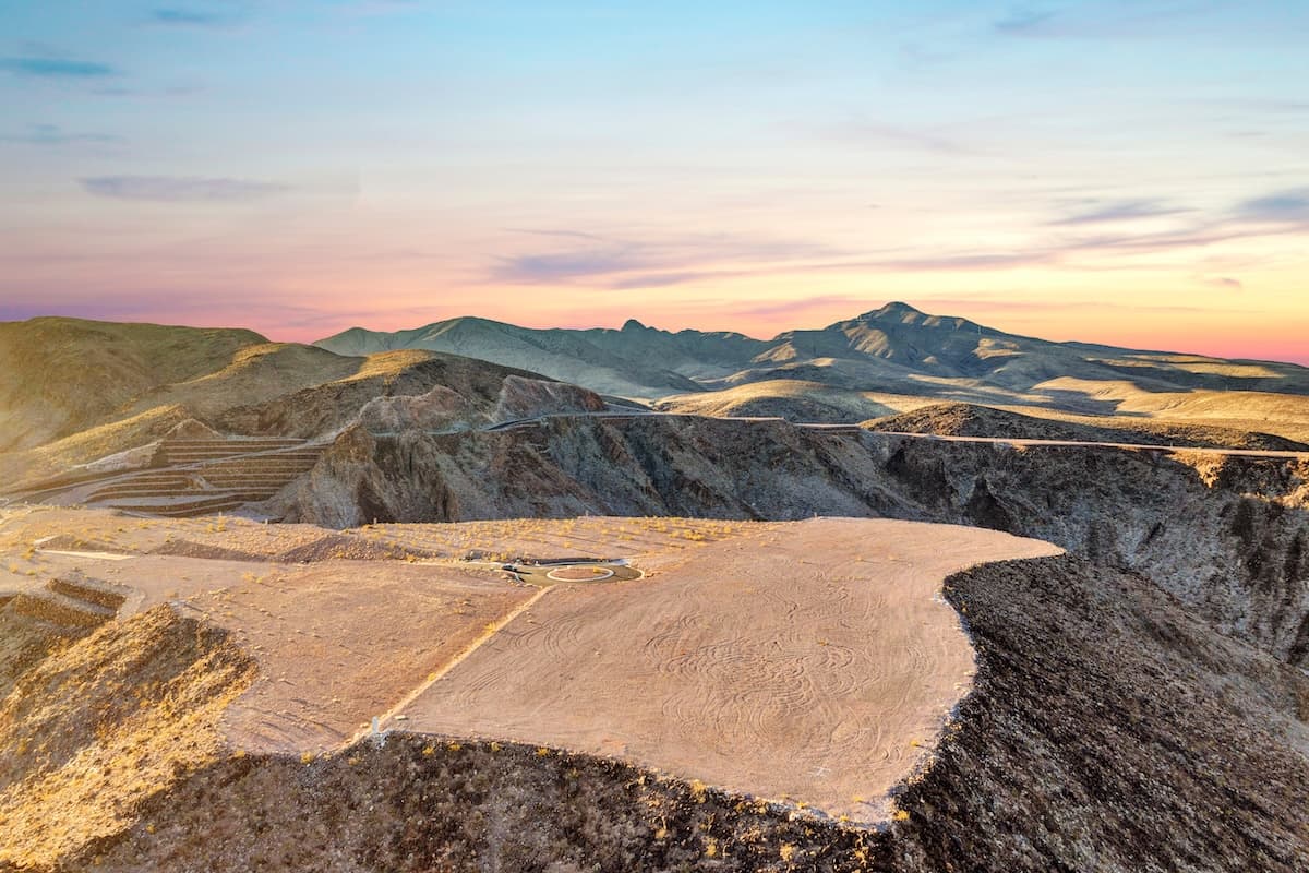 Aerial view of a desert hillside lot with a circular driveway pad overlooking a sprawling suburban city at sunset, with mountains in the distance.