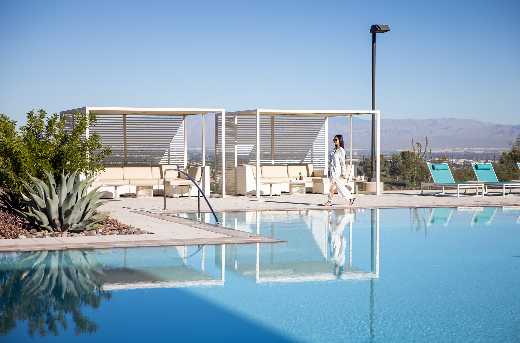 A resident walking alongside a serene zero-edge pool with private cabanas, lounge seating, and expansive views of the valley and surrounding mountains.