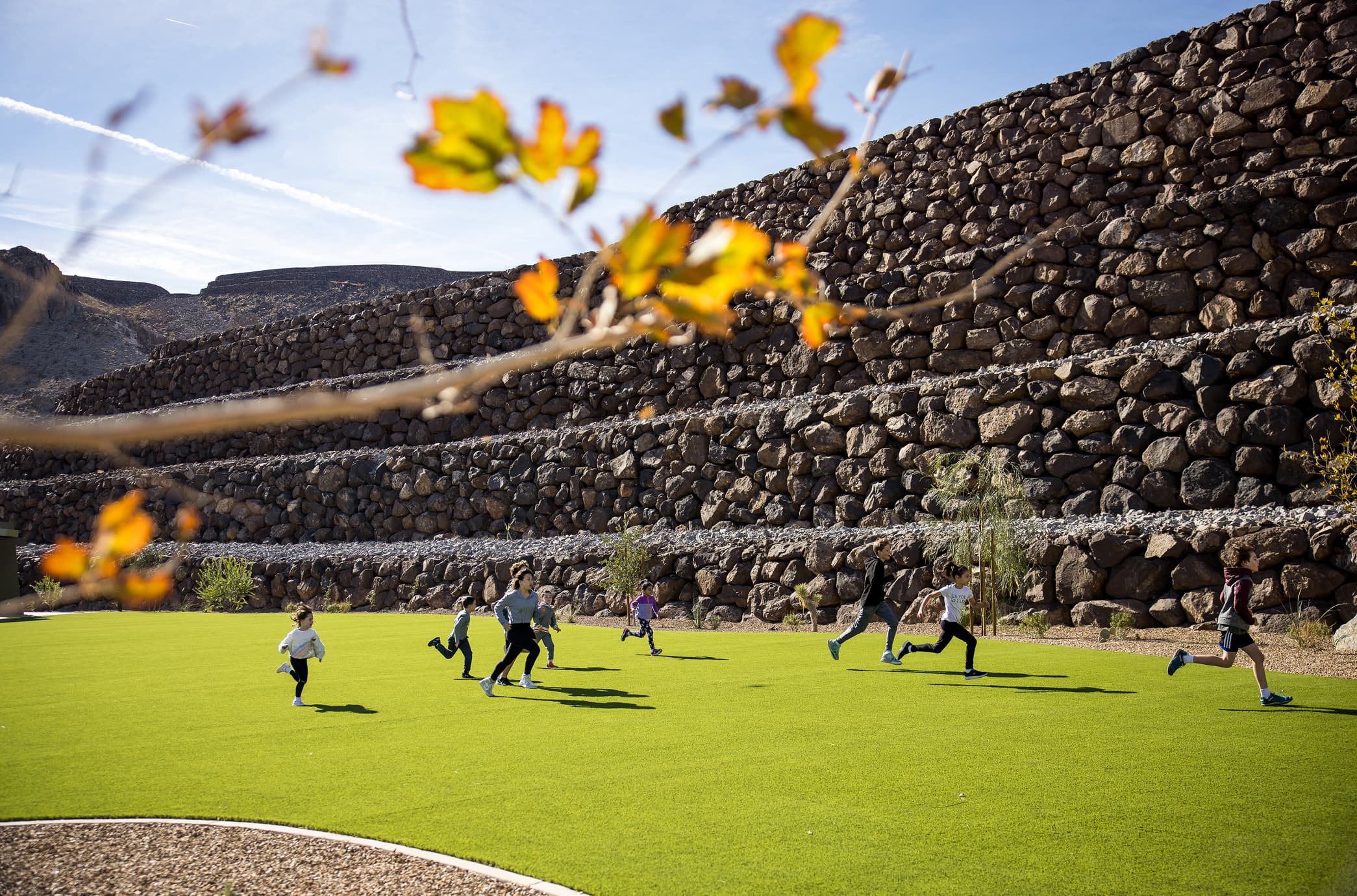 Children running and playing on a lush green lawn beneath terraced stone walls, set within a scenic desert landscape.