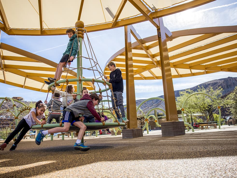 Children running and playing on a lush green lawn beneath terraced stone walls, set within a scenic desert landscape.