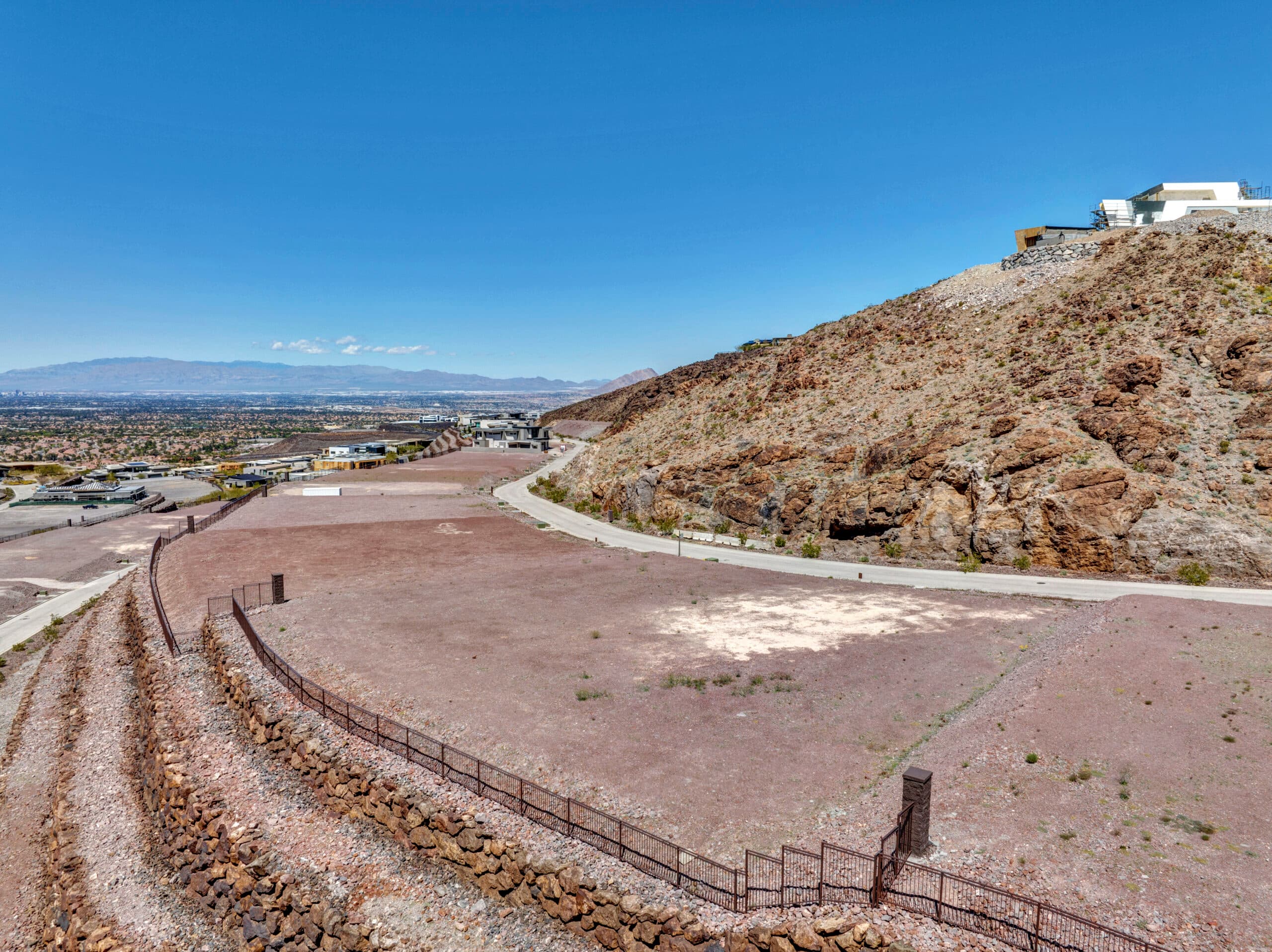 Desert hillside residential lot with mountain views, paved road access, and nearby modern homes under clear blue sky.