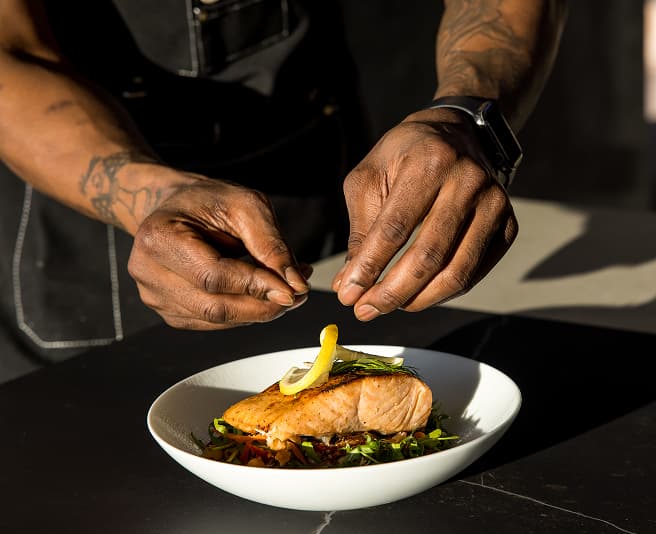 Chef plating gourmet salmon dish with lemon garnish and fresh greens in upscale restaurant setting.