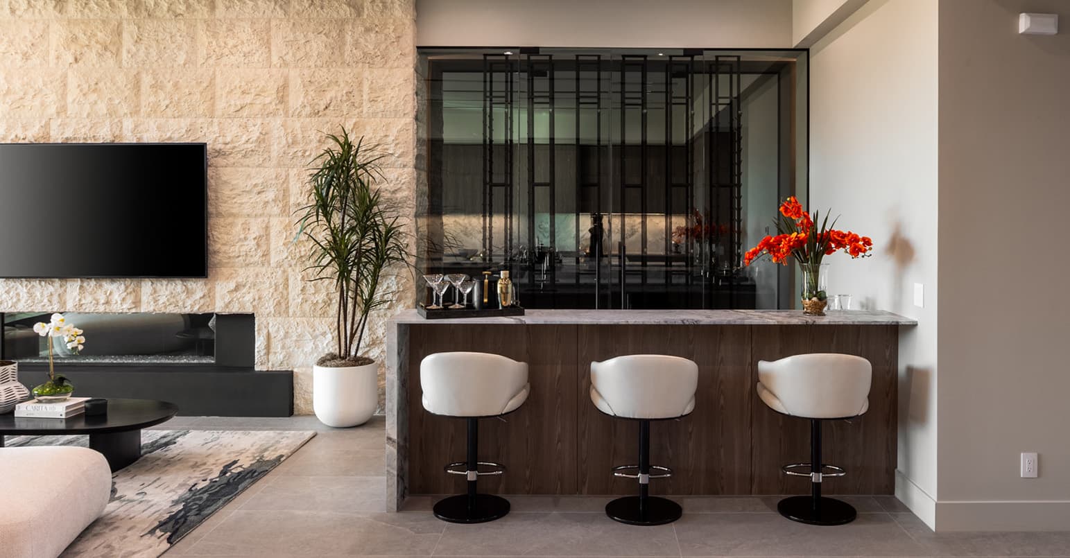 Modern indoor bar area with a sleek wood counter and three upholstered bar stools, backed by a glass-enclosed wine display, alongside a stone accent wall with a built-in fireplace and contemporary decor.