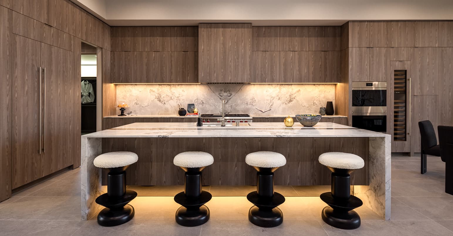 Contemporary chef’s kitchen with warm wood cabinetry, a waterfall marble island, integrated appliances, and sculptural bar seating, accented by soft under-cabinet lighting.