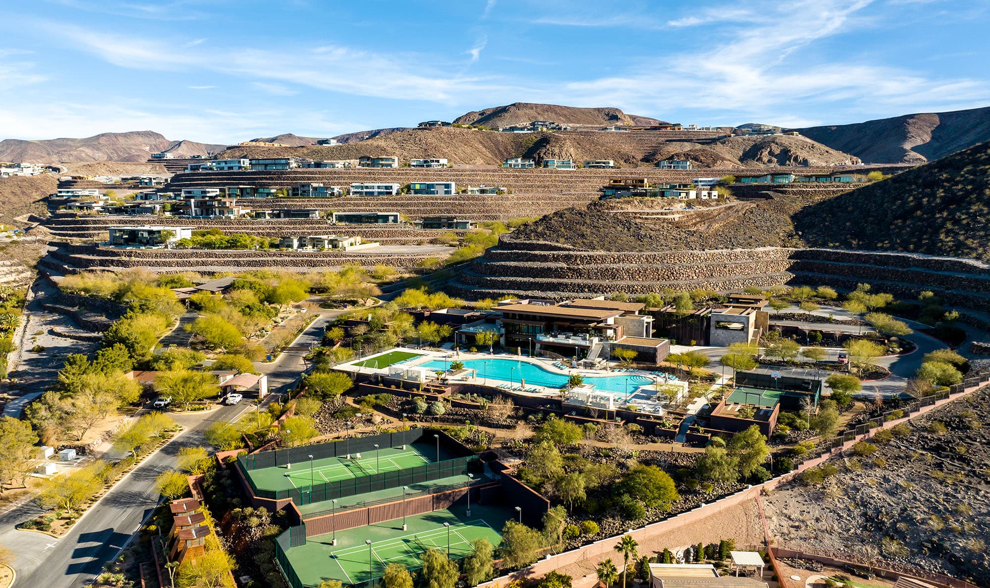 Aerial view of Ascaya’s terraced hillside community, showcasing modern luxury homes, the clubhouse, resort-style pool, tennis courts, and winding roads set against a dramatic desert landscape.
