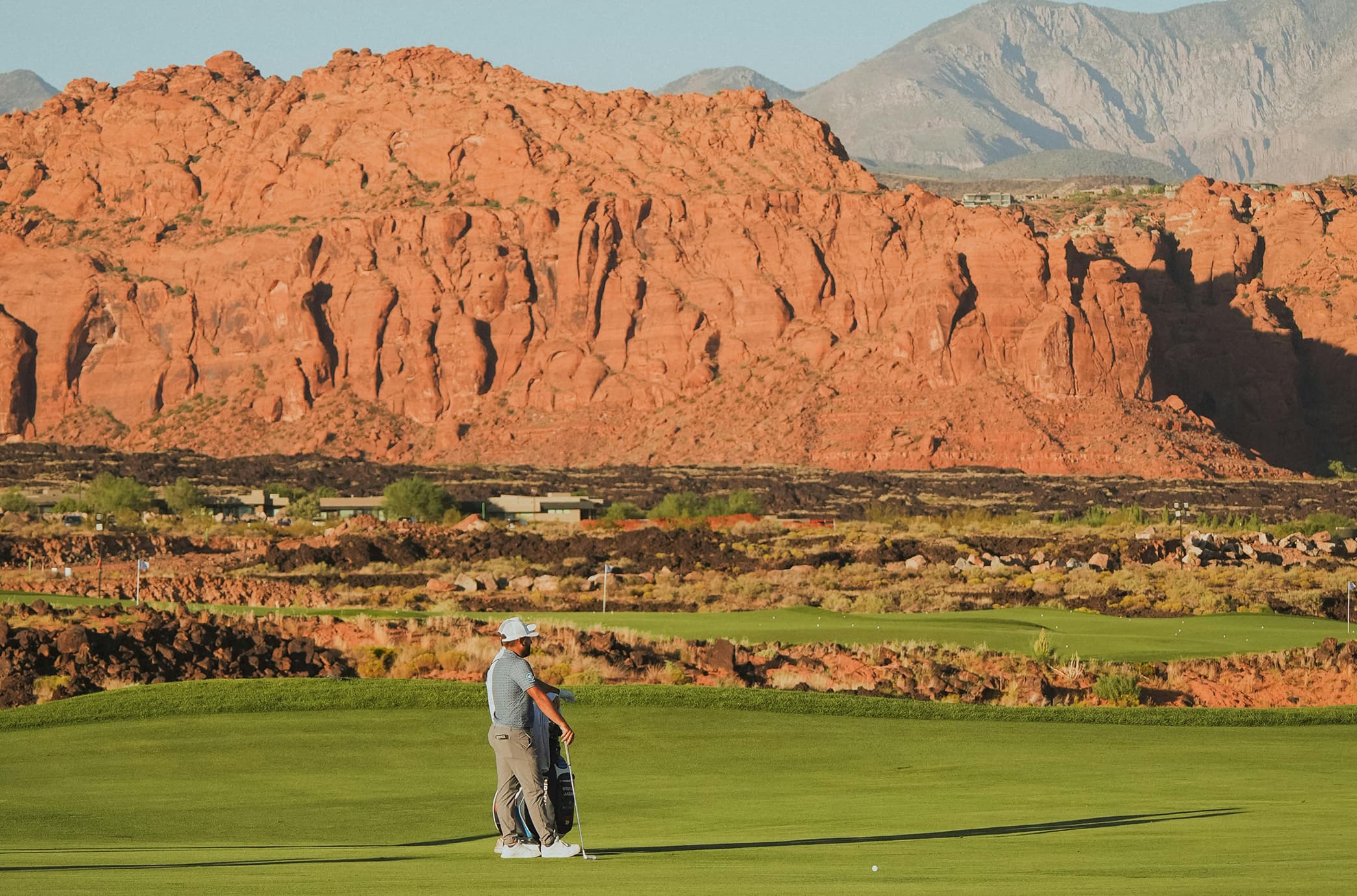 A golfer on a lush green fairway set against striking red rock cliffs and desert mountain scenery.