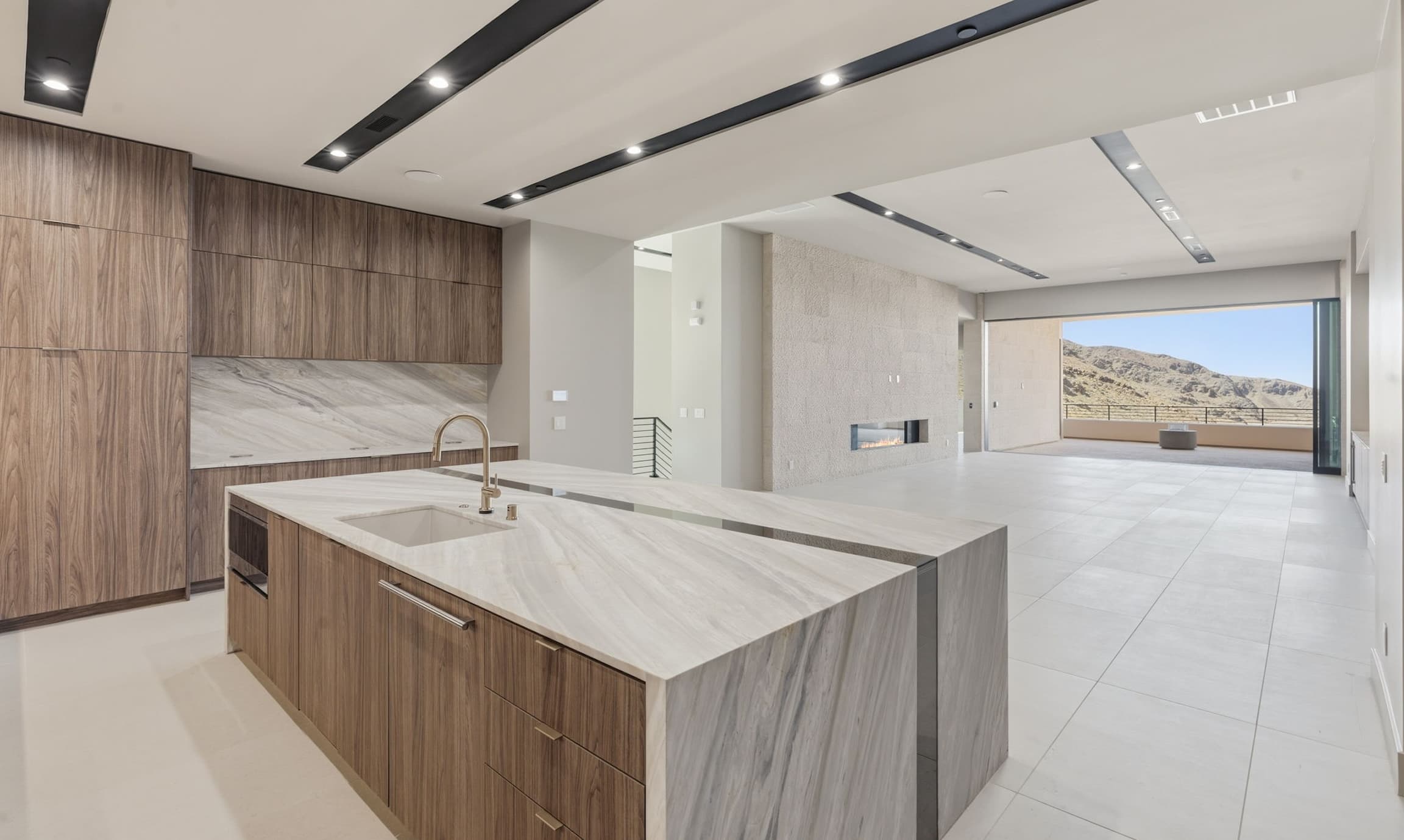 Modern open-concept kitchen and great room with a large waterfall-edge marble island, walnut cabinetry, recessed linear ceiling lights, and light tile flooring. The spacious interior features a sleek built-in fireplace and expansive sliding glass doors opening to a covered patio with mountain desert views.