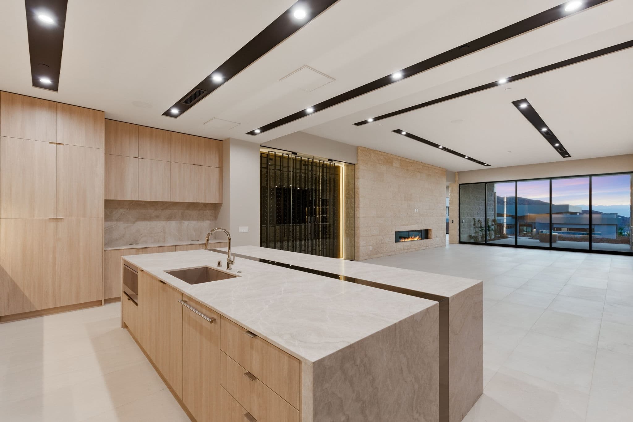 Open-concept great room featuring a chef’s kitchen, wine display, linear fireplace, floor-to-ceiling windows, and seamless indoor-outdoor living