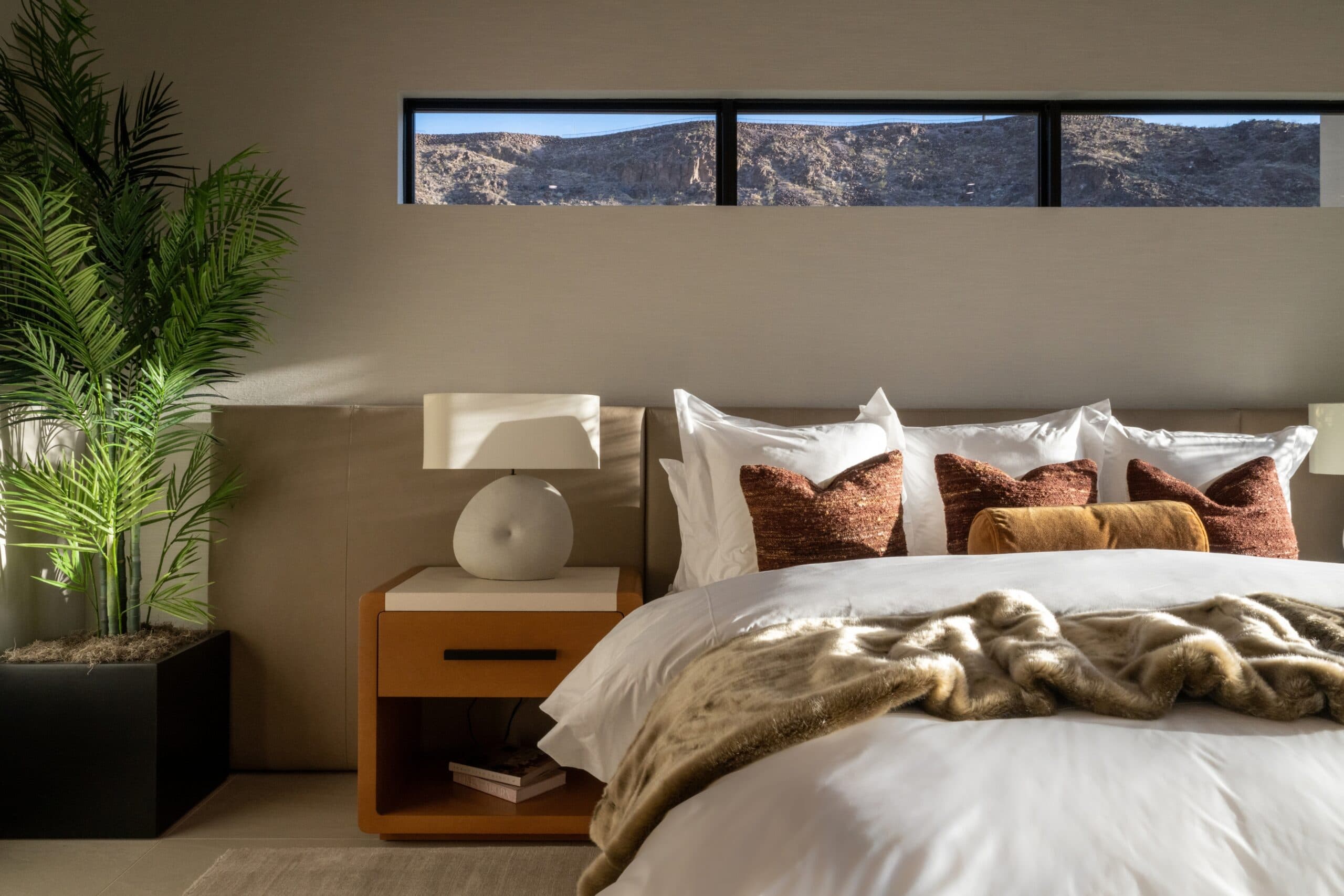 Luxury bedroom with layered neutral bedding, warm wood accents, greenery, and scenic desert mountain views.