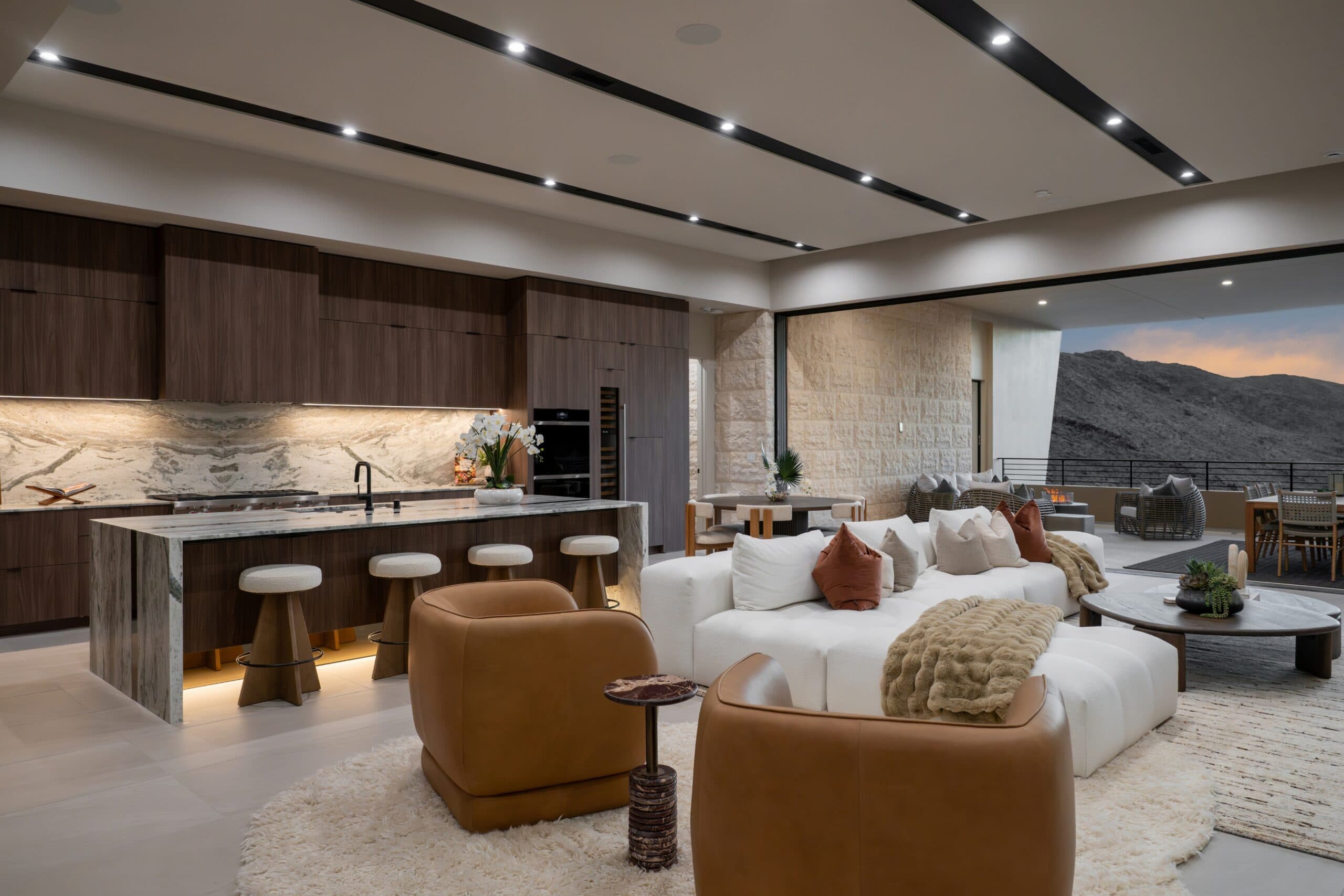 Luxury open-concept kitchen and living room with rich wood cabinetry, marble finishes, designer furnishings, and expansive desert mountain views.