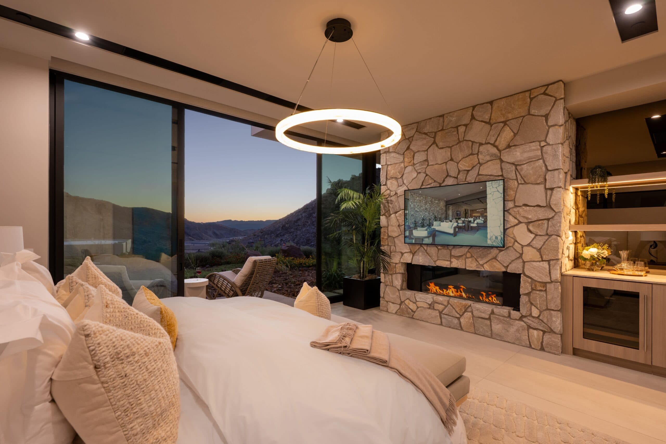 Luxury modern bedroom with a stone fireplace, floor-to-ceiling windows, and serene desert mountain views at sunset.