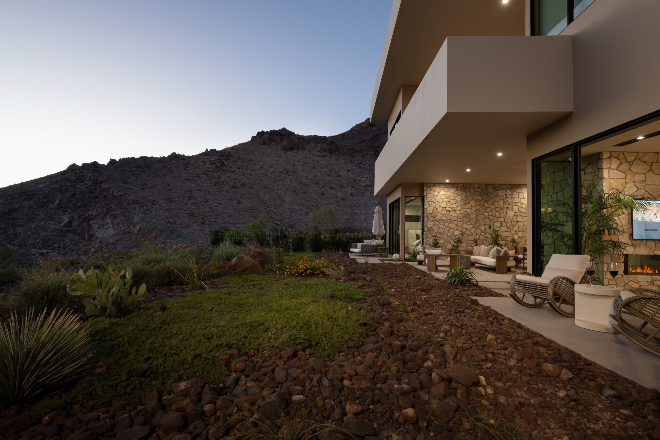 Luxury modern desert home with covered outdoor seating, stone accents, lush landscaping, and dramatic mountain views at dusk.