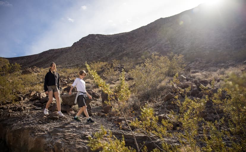 Two people hike down a rocky desert trail under bright sunlight.