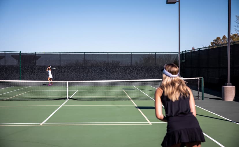 Two women play tennis on an outdoor court under a clear blue sky.