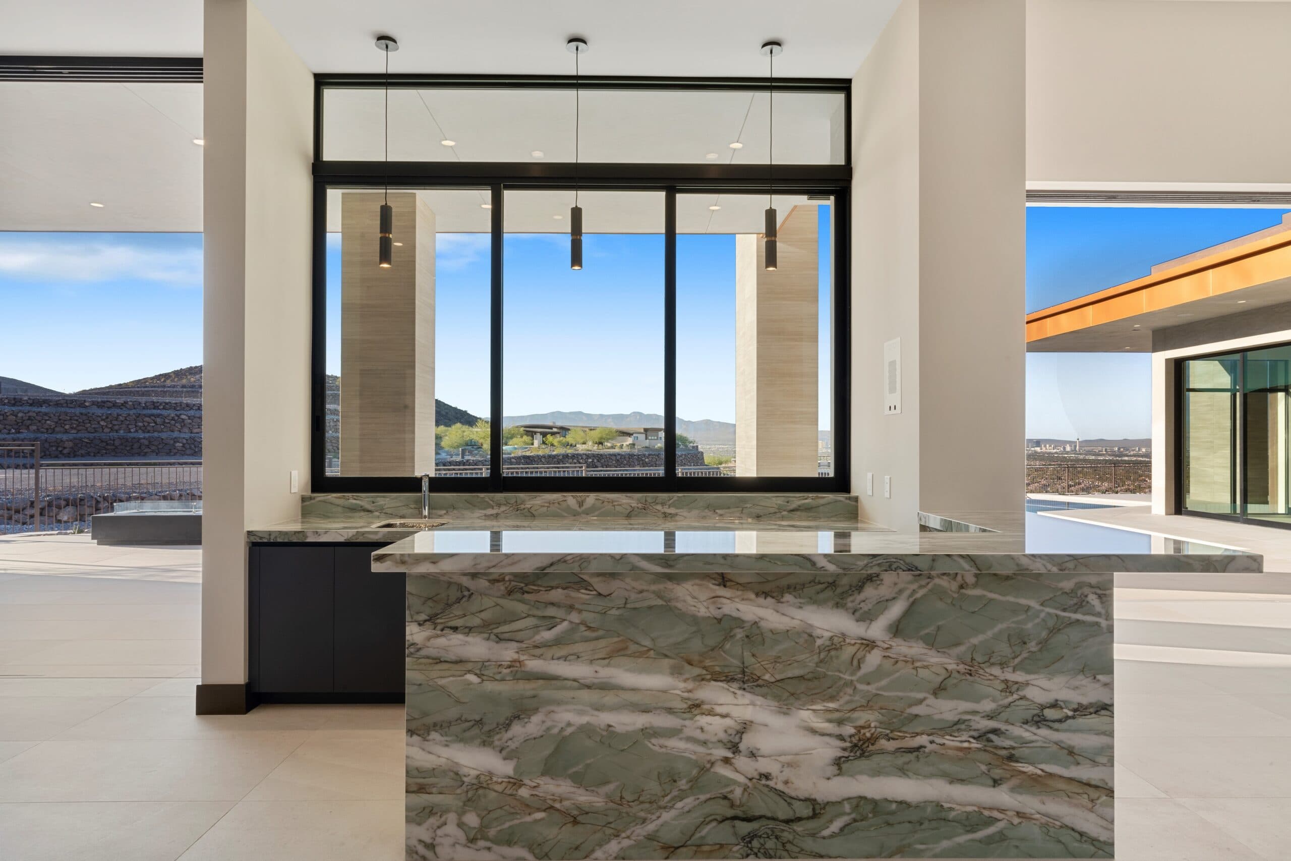 Modern luxury kitchen featuring a dramatic marble waterfall island, floor-to-ceiling windows with desert and mountain views, and seamless indoor-outdoor living with contemporary architectural design.