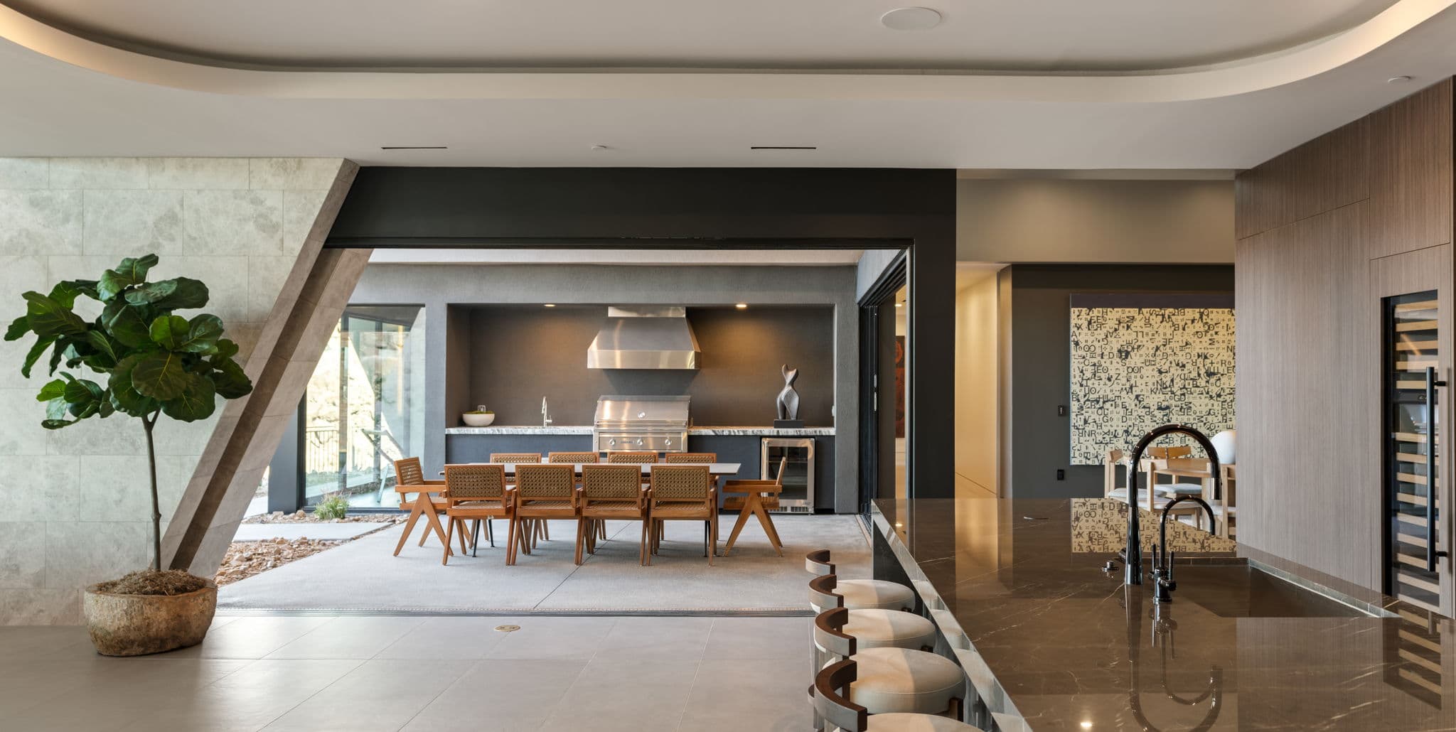 Modern luxury kitchen with a sleek dark stone island, integrated wine storage, and seamless indoor-outdoor living that opens to a covered outdoor dining area with a built-in grill and contemporary desert-inspired design.